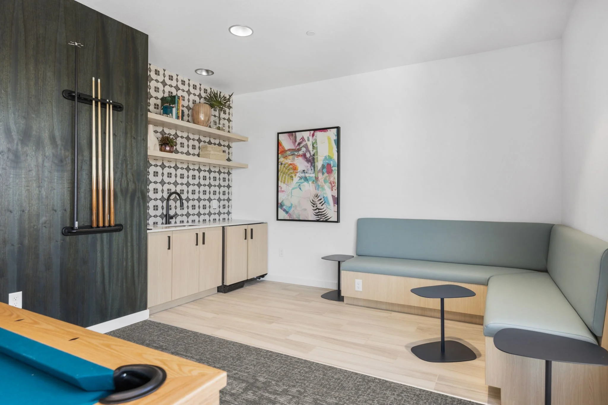 Modern game room with light wood flooring, pool table, navy blue and light green built-in seating, abstract art on white wall, open shelving with decorative items, and a black cue holder on the wall.