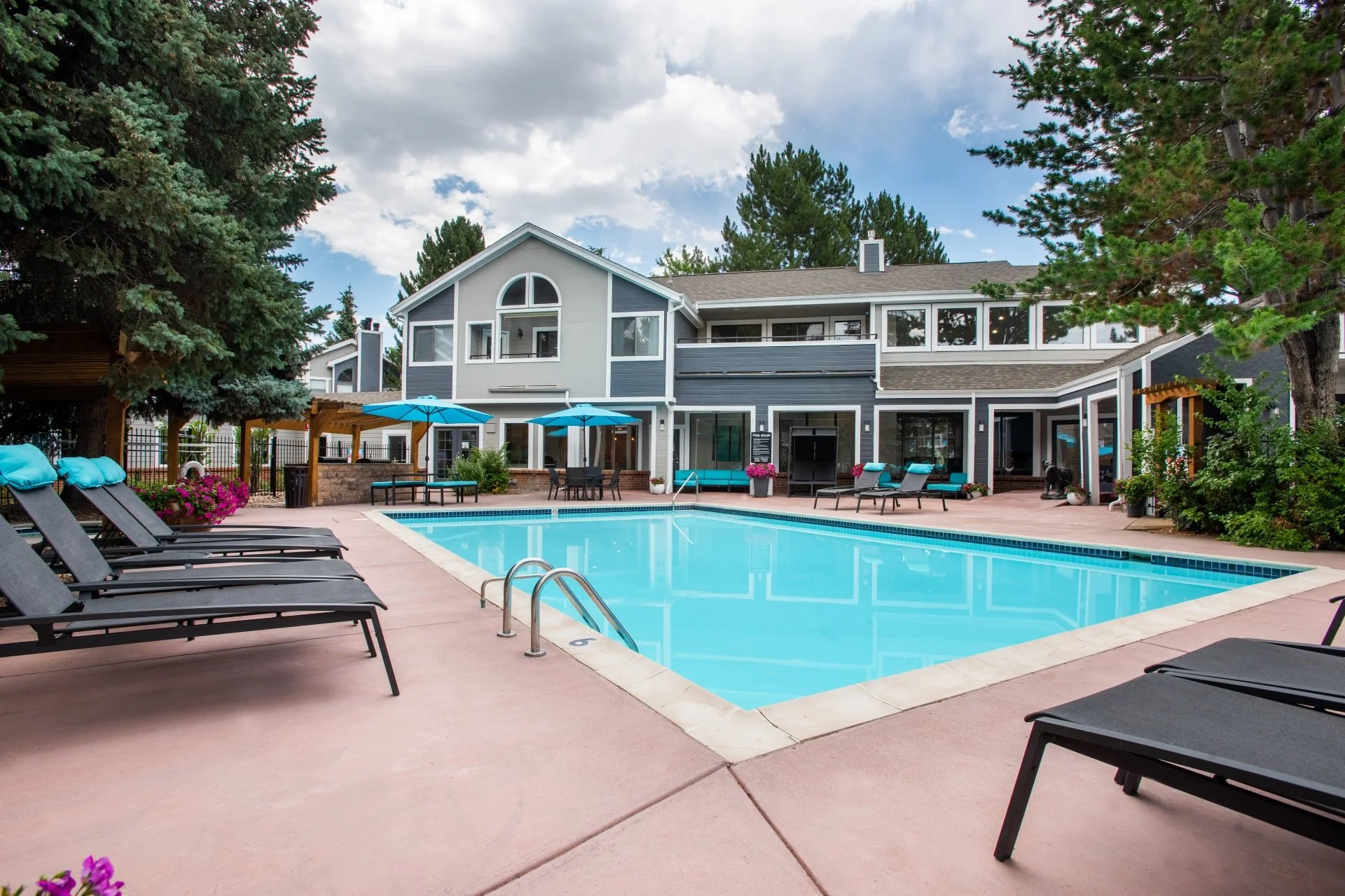 Swimming pool surrounded by lounge chairs and umbrellas in a residential backyard, with a multi-story house and tall trees in the background.