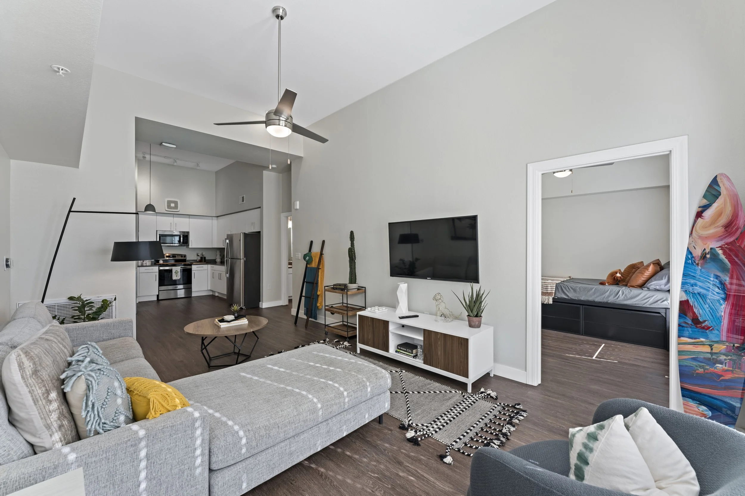Living room with gray sectional sofa, small round coffee table, wall-mounted TV, modern white TV stand, black and white patterned rug, kitchenette in the background, and a doorway leading to a bedroom with a bed and colorful art piece.