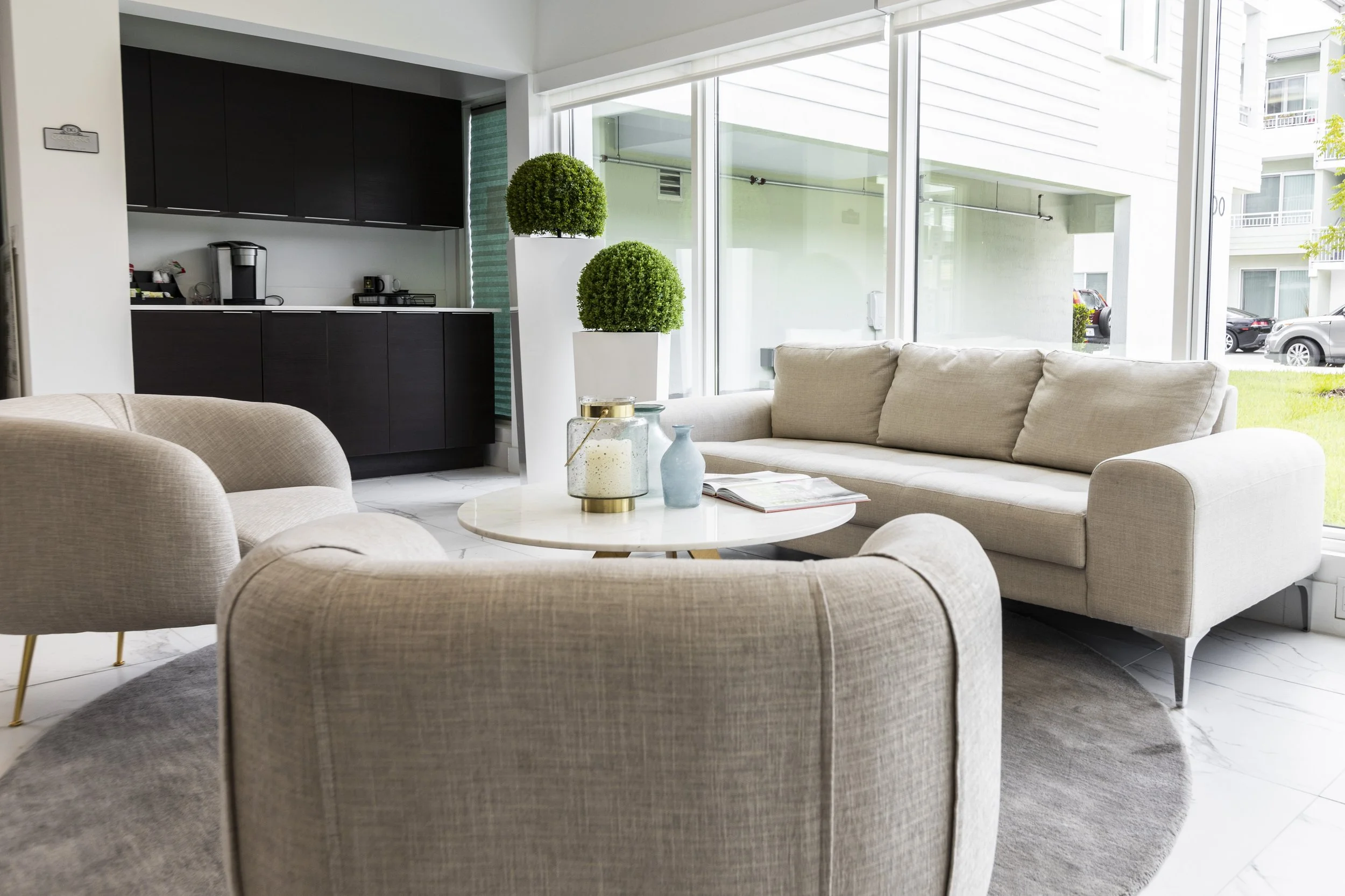 Modern living room with beige sofas, a round coffee table, decorative vases, and high glass windows showing a grassy lawn outside.