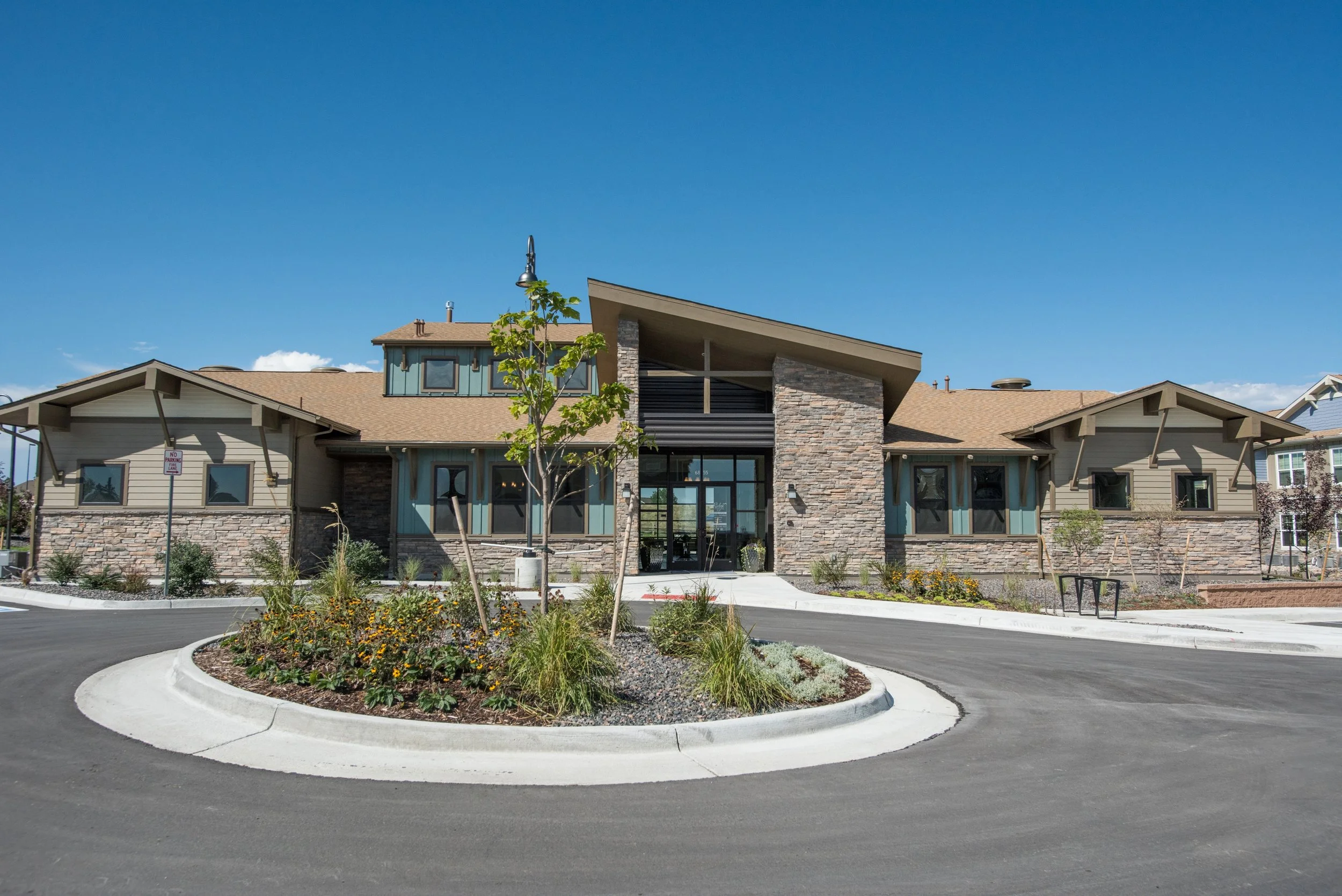 A modern, multi-story residential building with a mix of stone and beige siding, large glass entryway, and a sloped roof under a blue sky.