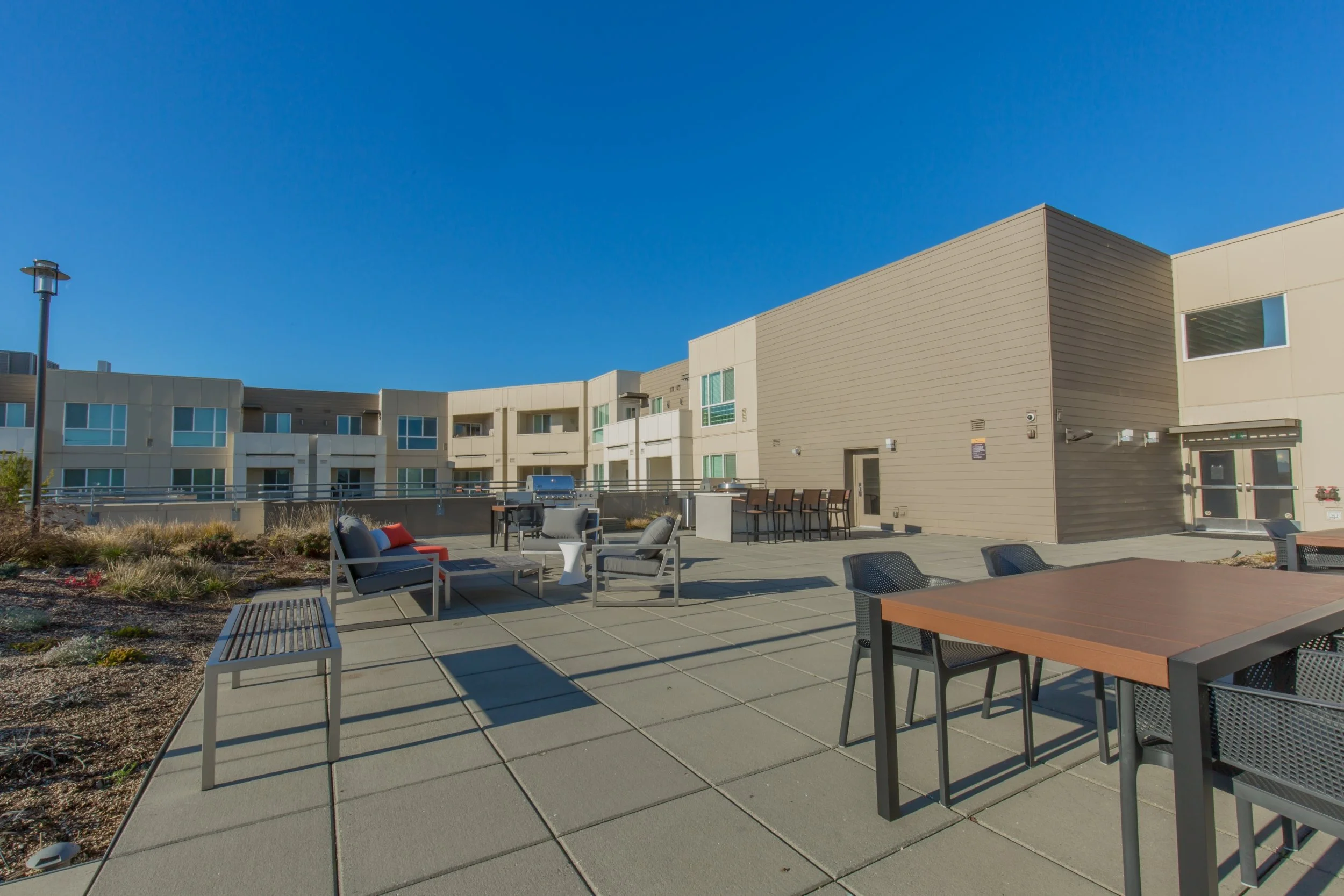 Outdoor patio with seating and grilling area in front of modern apartment complex under a clear blue sky.