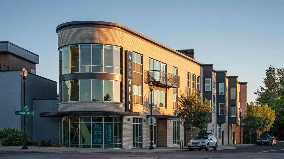 Modern four-story apartment building with rounded corner and large glass windows, sign reads "ATTWELL," located at the intersection of Burnham Street and another street, with cars parked outside and trees nearby.