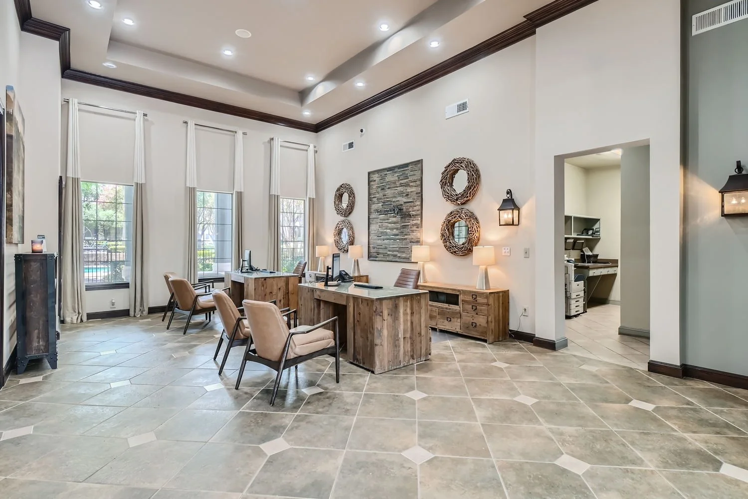A spacious lobby area with beige chairs, wooden desks, and large windows with white curtains, featuring decorative wall art and lighting fixtures.