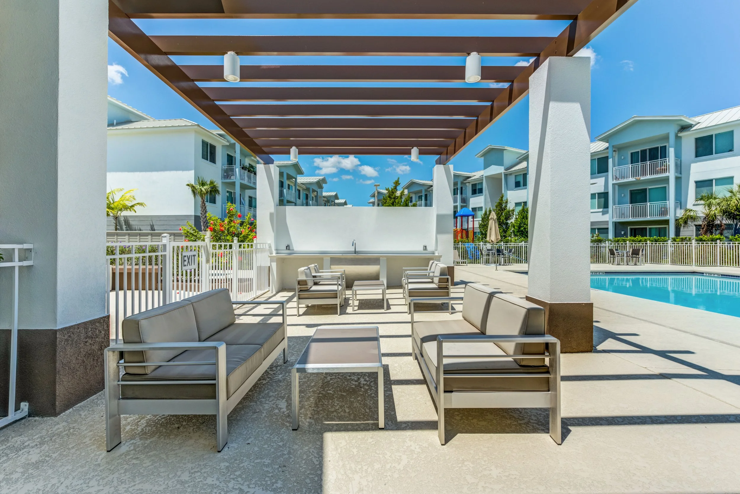 Poolside sitting area with beige cushioned outdoor furniture, a small table, a pool, and surrounding multi-story apartment buildings with balconies under a clear blue sky.