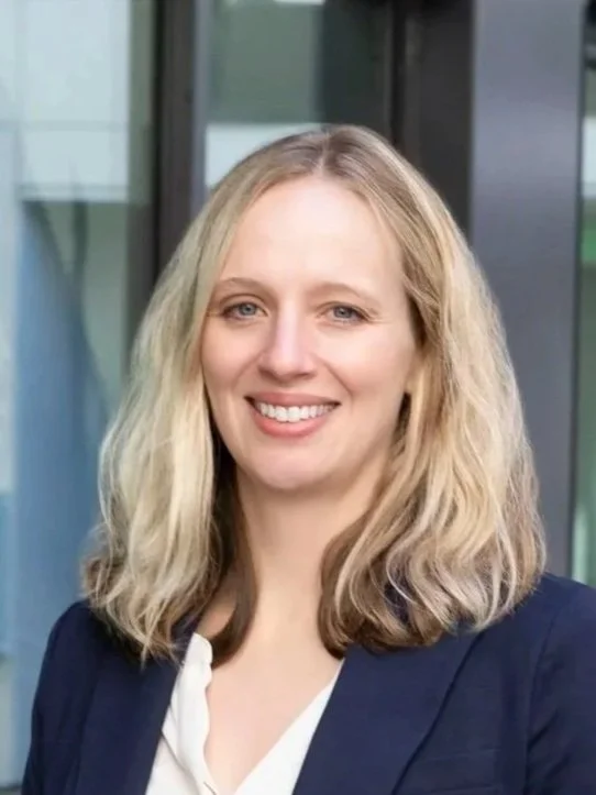 A smiling woman with long blonde hair, wearing glasses with a dark frame and a black blazer, against a blue background.
