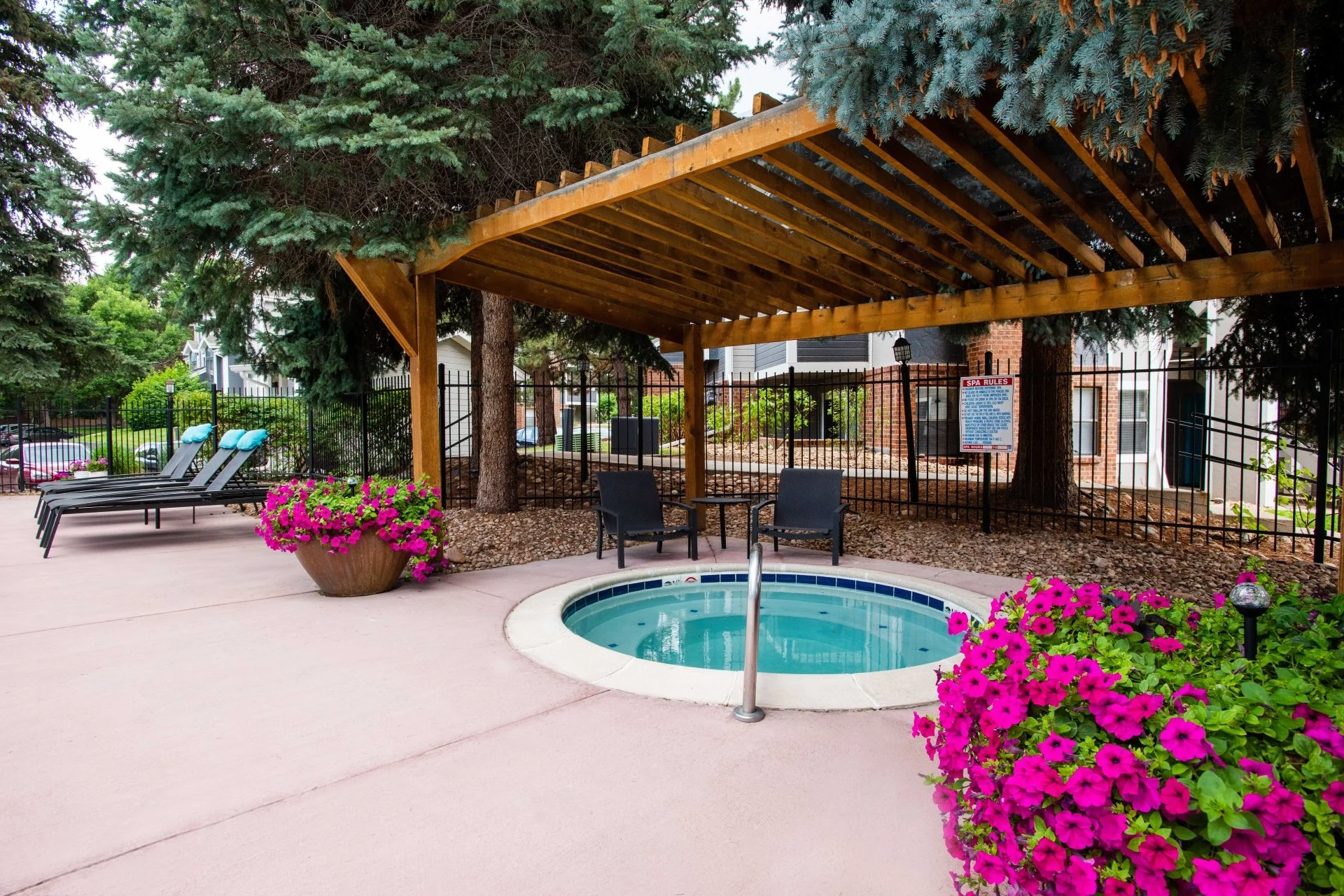 An outdoor pool area with lounge chairs, a hot tub, pink flowers in pots, and shaded seating under a wooden pergola, surrounded by trees and fences.