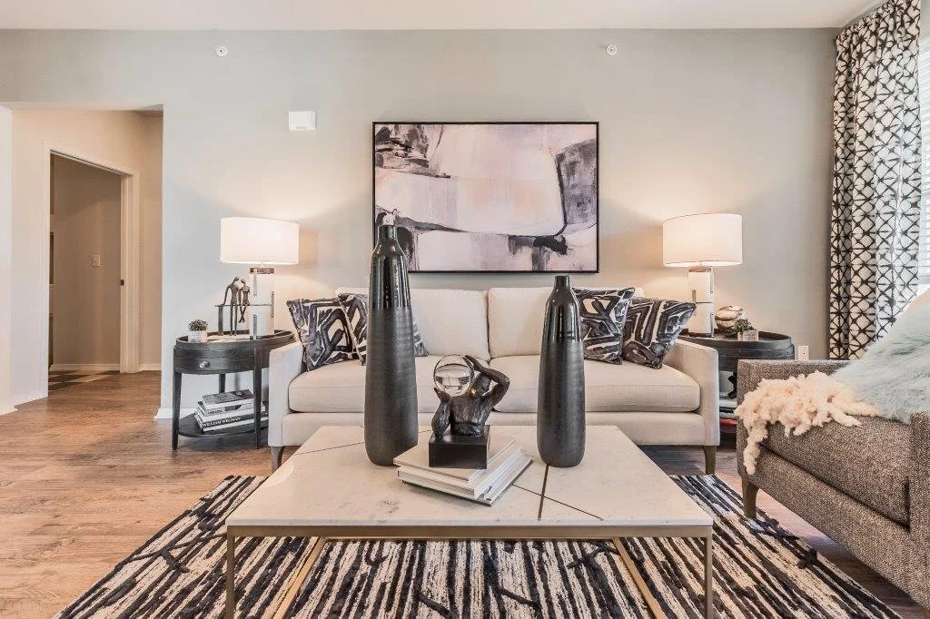 Living room with a beige sofa, black and gray decorative pillows, wood coffee table with black vases and decorative objects, two side tables with lamps, artwork on the wall, patterned curtains, and a gray armchair with a throw blanket.
