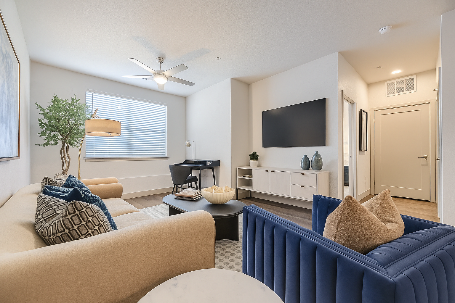 Modern living room with beige and navy blue sofas, decorative pillows, a coffee table with a bowl and books, a flat-screen TV on a white TV stand, a small desk with a chair near a window, and a ceiling fan.