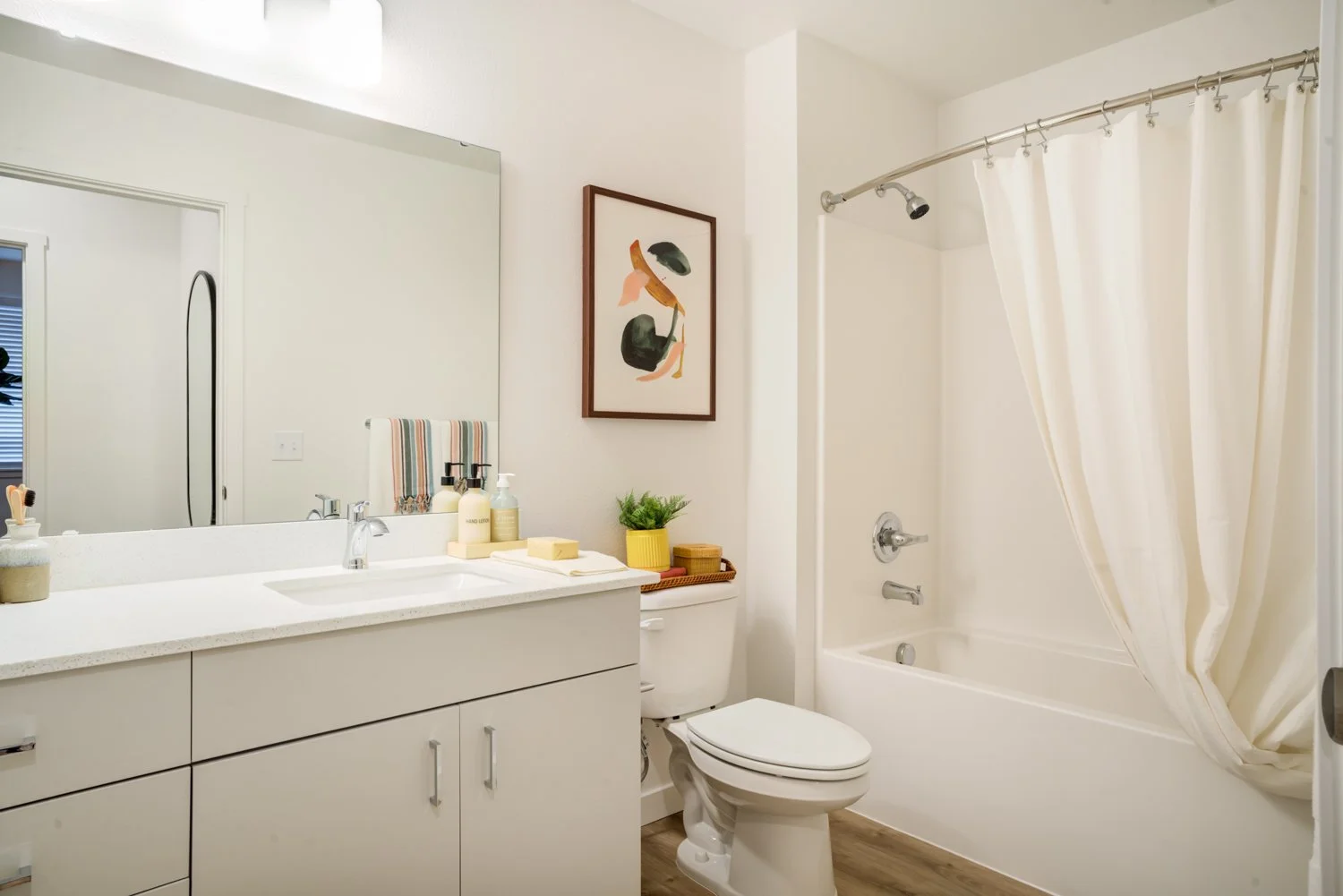 Bright bathroom with a large mirror above a white vanity, a framed abstract art on the wall, a toilet with a small plant and decorative items on the tank, and a bathtub with a cream-colored shower curtain.