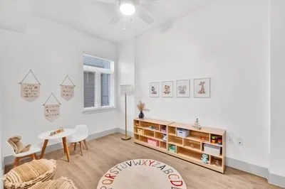 A bright children's playroom with white walls, a wooden floor, and a round colorful alphabet rug. There is a wooden shelf filled with books and toys, a window with blinds, and wall decorations including hanging fabric signs and framed pictures of ani