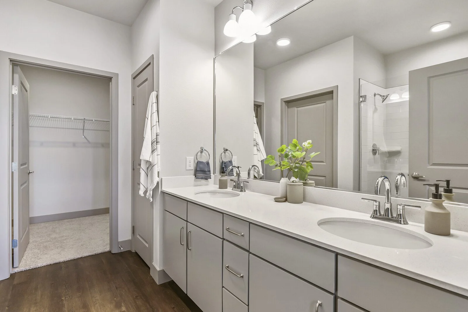 A modern bathroom with a double vanity, large mirror, potted plant, and white countertop. There is a walk-in closet with a wire shelf and a shower with white tiles. Neutral colors and wood floor.