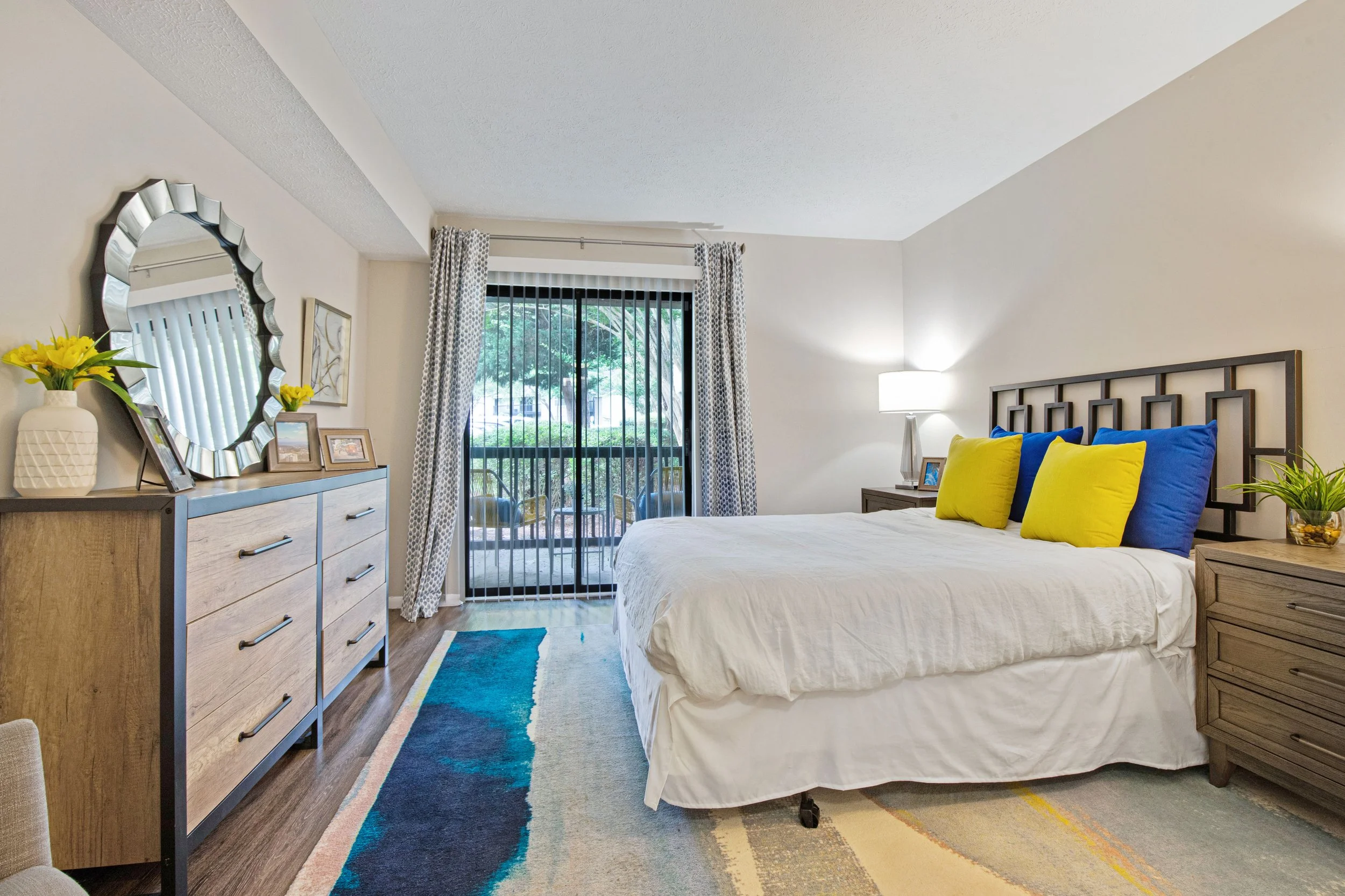 A bedroom with a large bed featuring blue and yellow pillows, a wooden dresser with a mirror, and a sliding glass door leading to a balcony or patio.