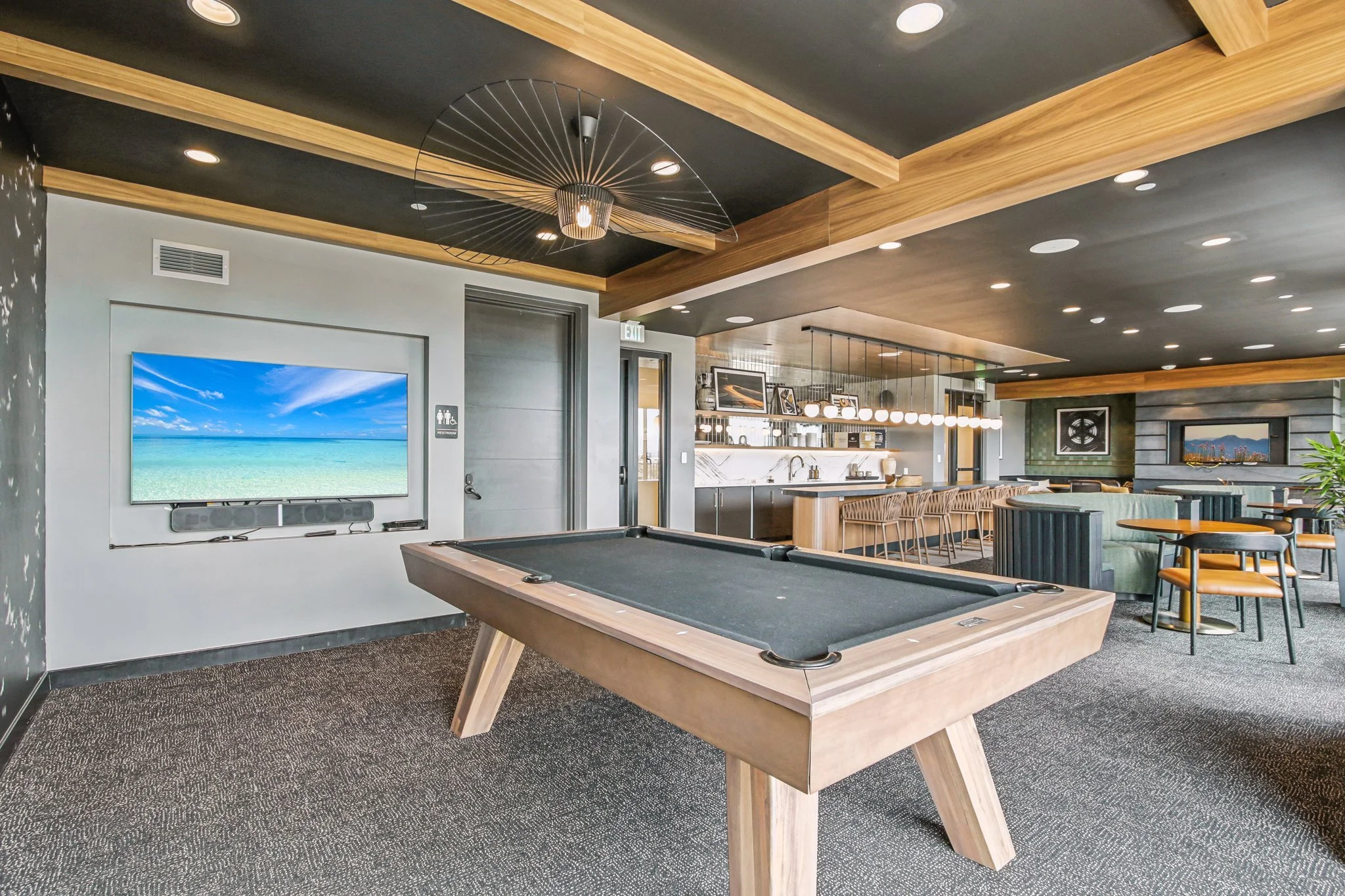 Interior of a modern lounge area with a pool table, large flat-screen TV displaying a beach scene, bar counter, and seating arrangements, including chairs and couches, with a well-lit ceiling and contemporary decor.