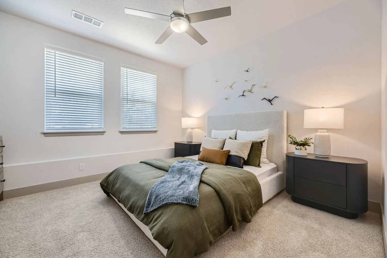 A bedroom with two windows having white blinds, a ceiling fan with a light, a bed with green bedding and pillows, two black nightstands with table lamps, and decorative birds on the wall.