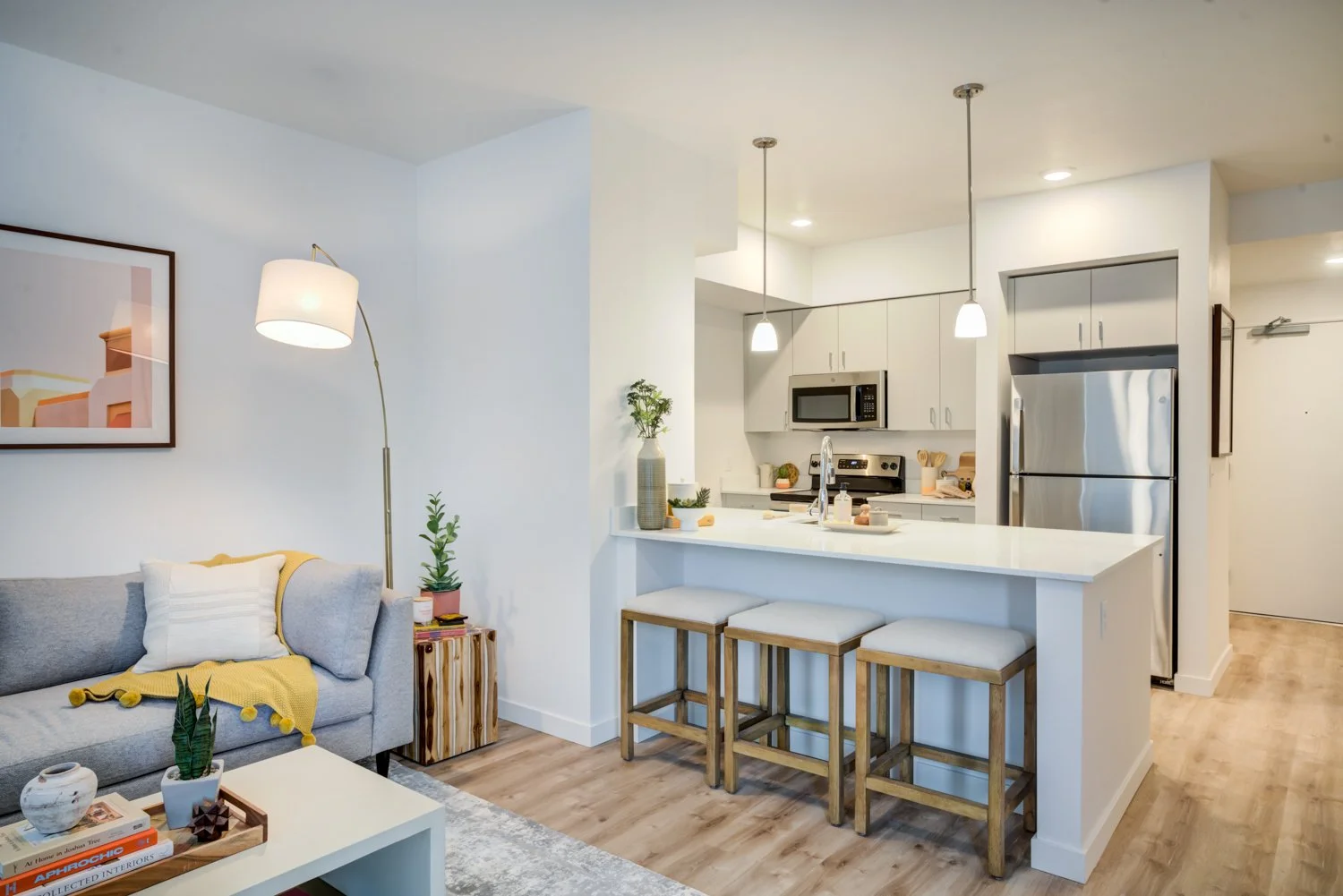 Modern open-concept living room and kitchen with white and gray decor, featuring a gray sofa with yellow throw, a white coffee table, a white kitchen island with three bar stools, stainless steel refrigerator, microwave, stove, pendant and recessed l