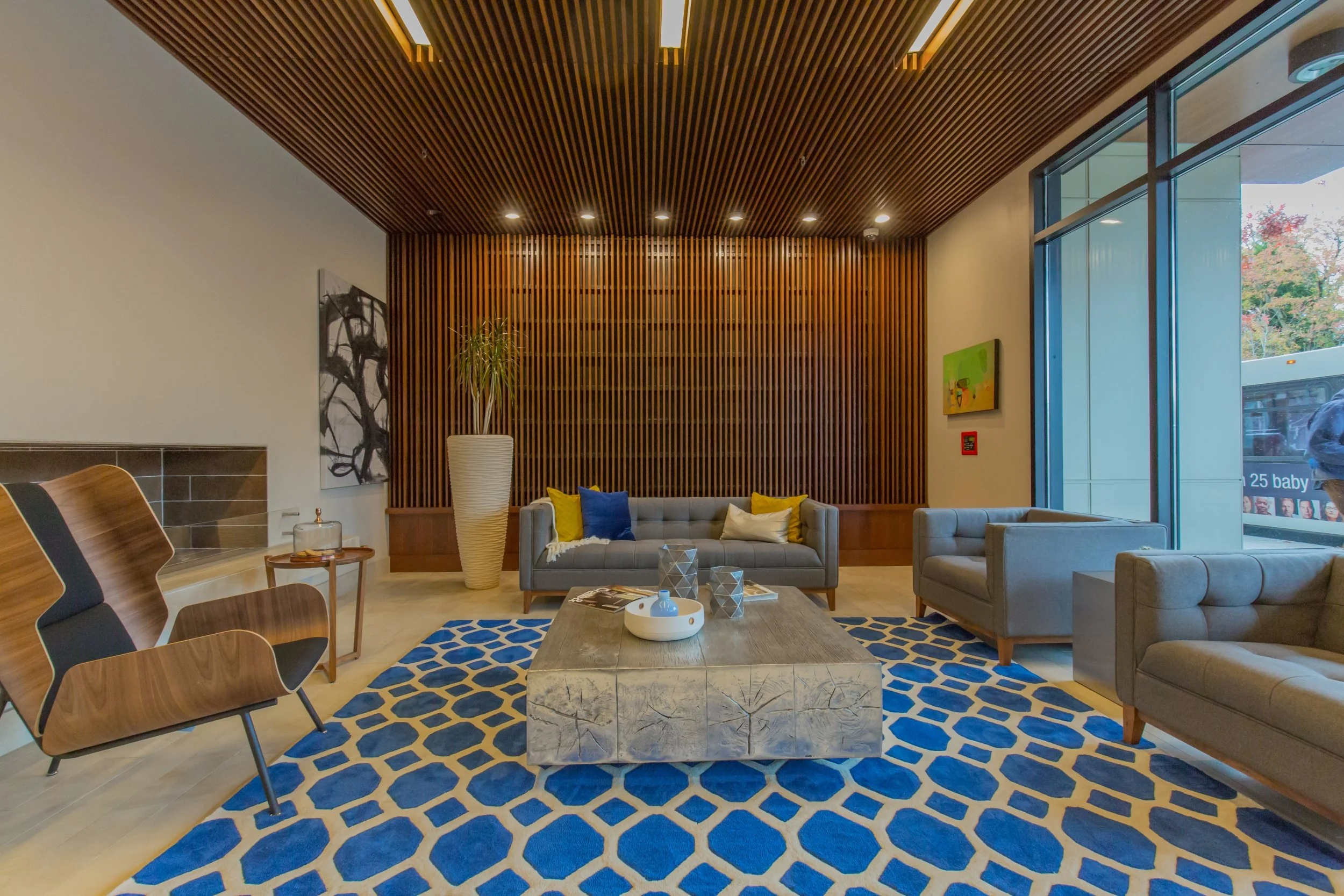 Modern hotel lobby with gray sofas, a wooden coffee table, blue and yellow cushions, a large patterned blue rug, and a wood-paneled wall background. Bright natural light from large window.