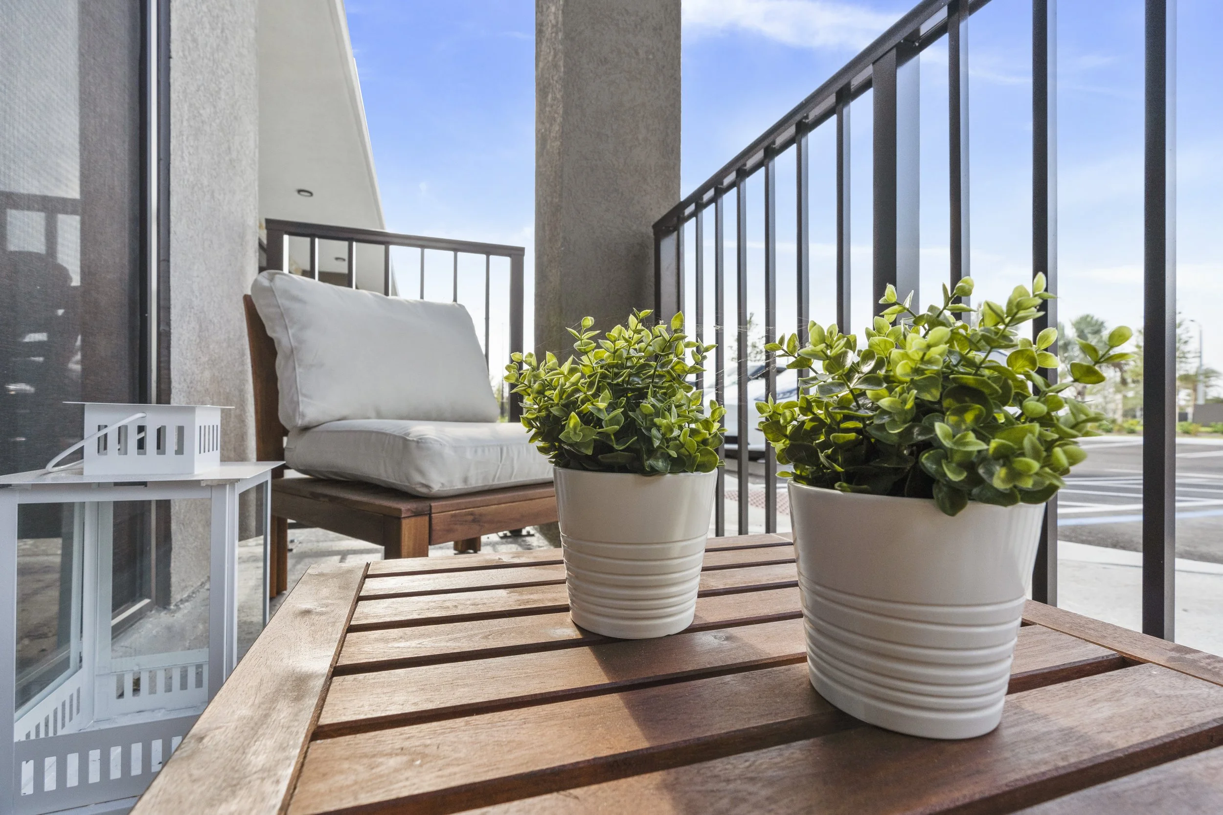 Two potted green plants on a wooden table on a balcony with a metal railing, outdoor furniture, and a blue sky with some clouds.