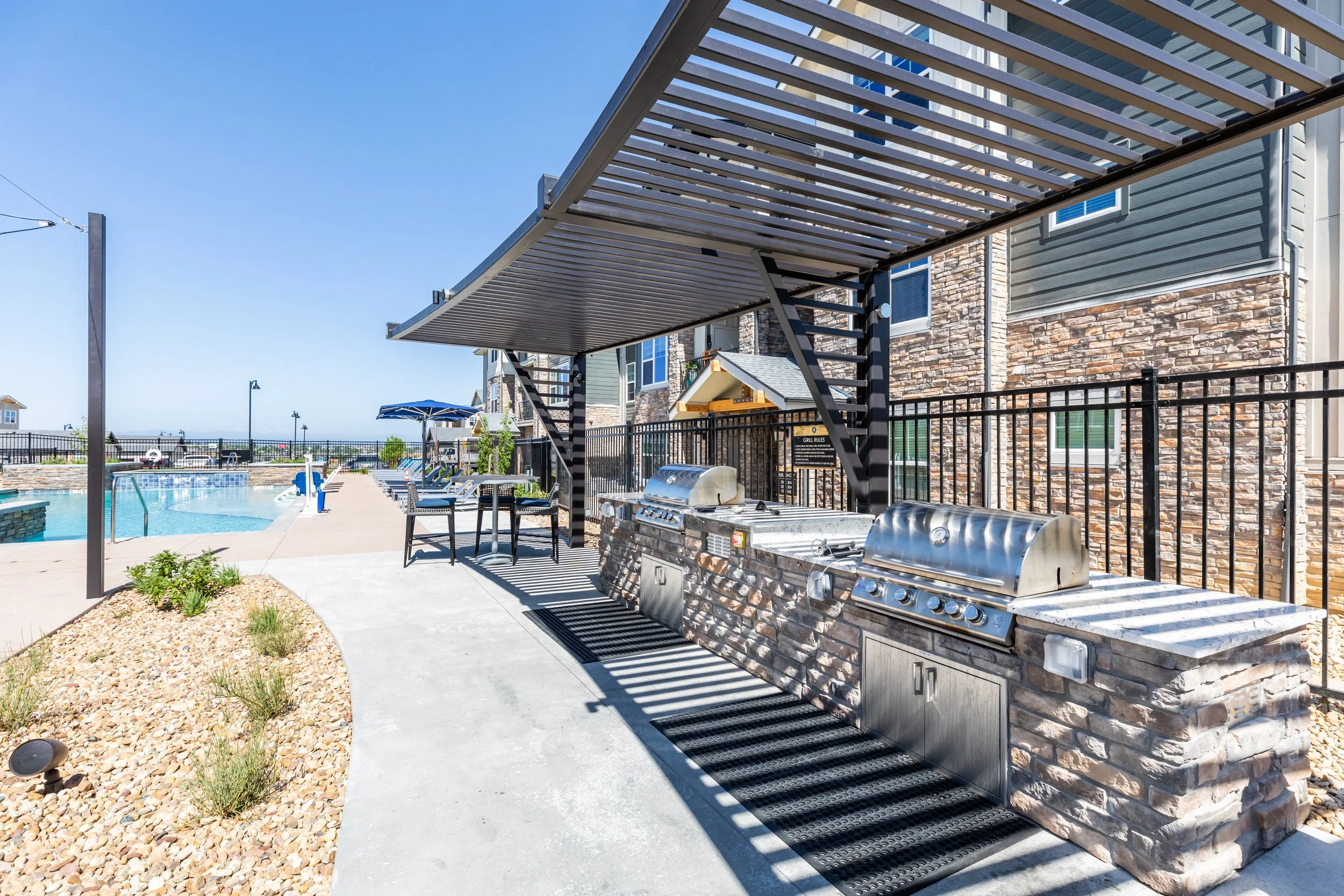 Outdoor swimming pool with lounge chairs and umbrellas, two stainless steel grills with a black metal pergola overhead, surrounded by a black fence, next to a multi-story residential building under a clear blue sky.