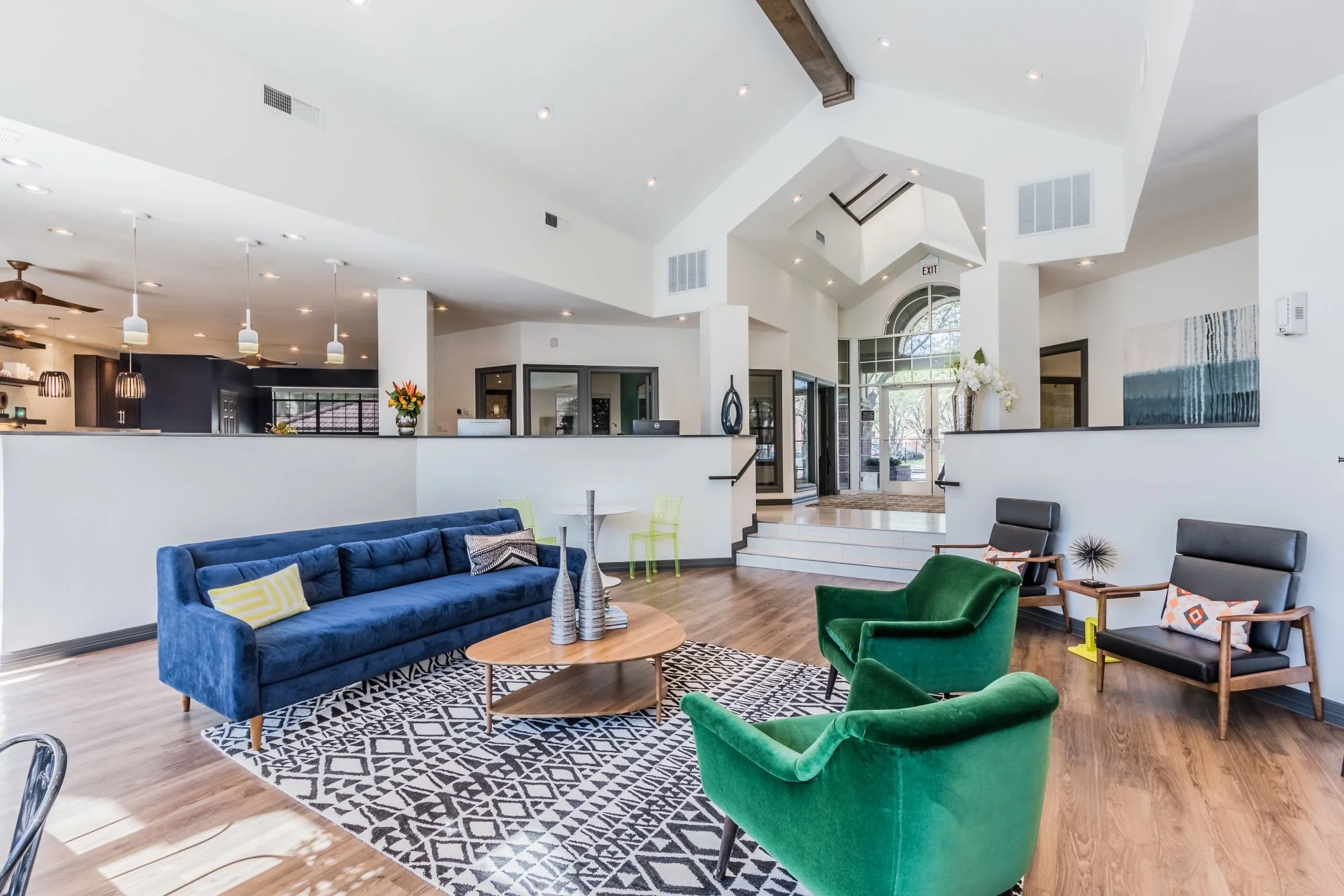 Modern hotel lobby with high vaulted ceilings, white walls, and hardwood floors, featuring a blue tufted sofa, green armchairs, black lounge chairs, a round wooden coffee table with decorative vases, a patterned rug, and fresh flower arrangements nea