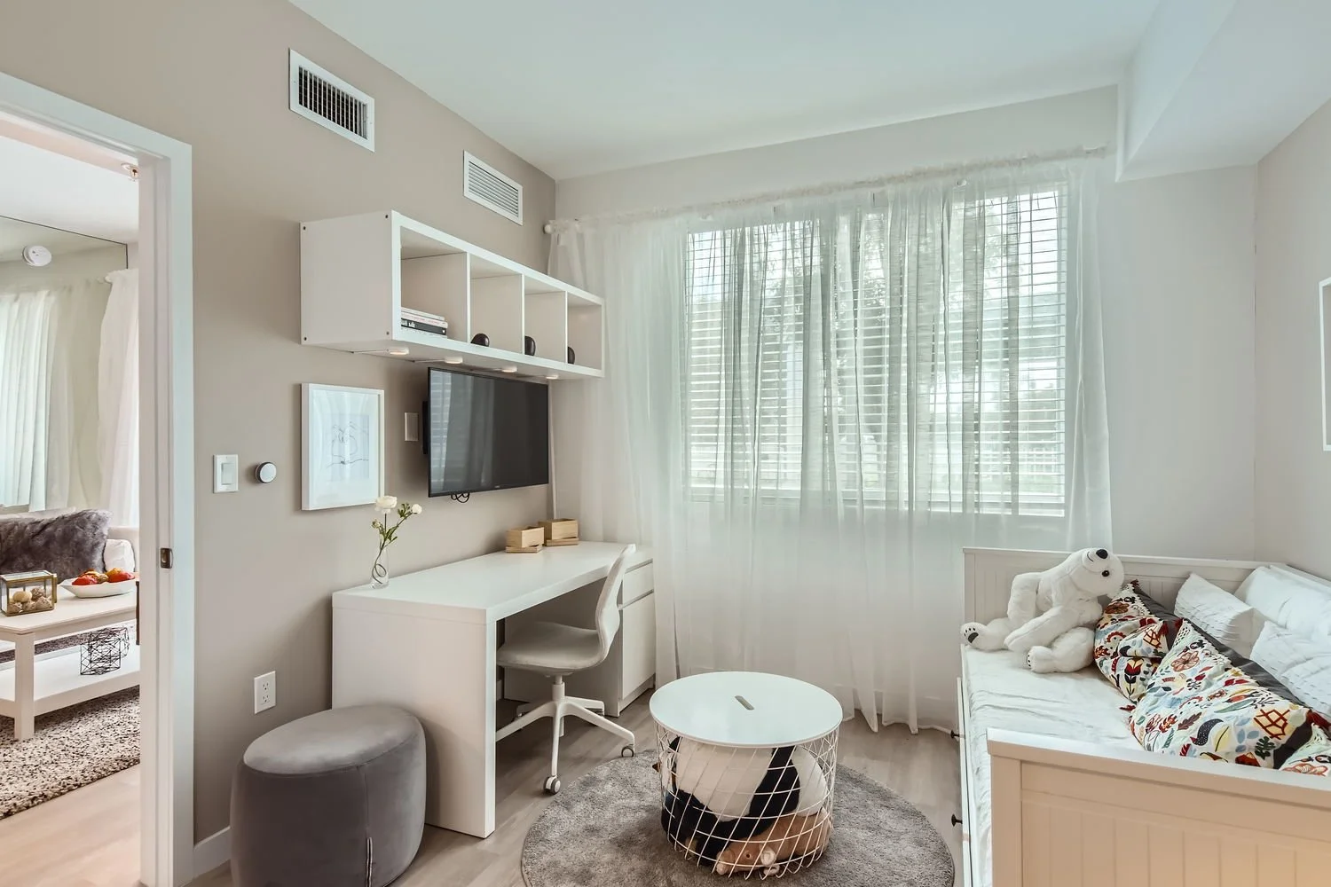 A bright, modern living room with a white desk, a gray ottoman, a small white round coffee table, a white daybed with decorative pillows and a plush white dog toy, a wall-mounted TV, white shelves, and large windows with sheer white curtains.