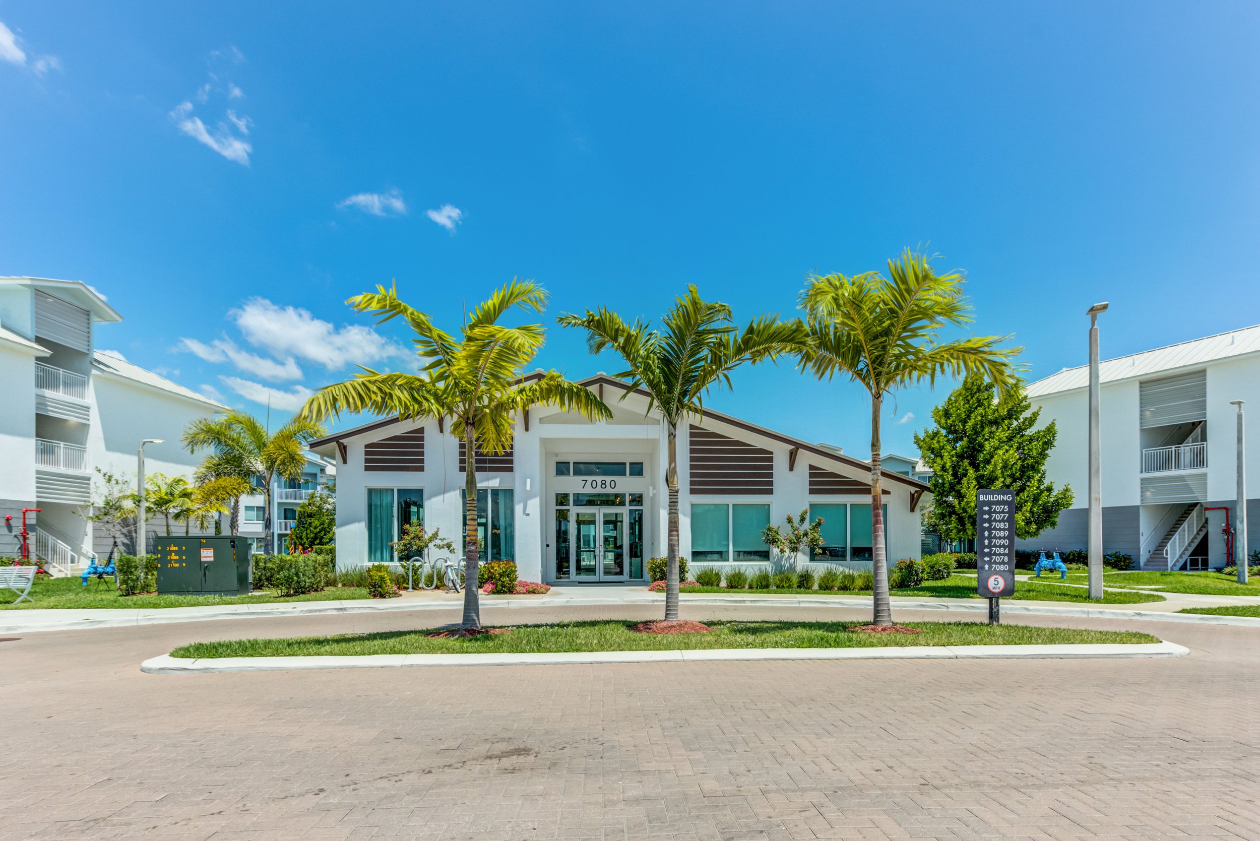 A modern residential building with palm trees in front, under a bright blue sky with some clouds.