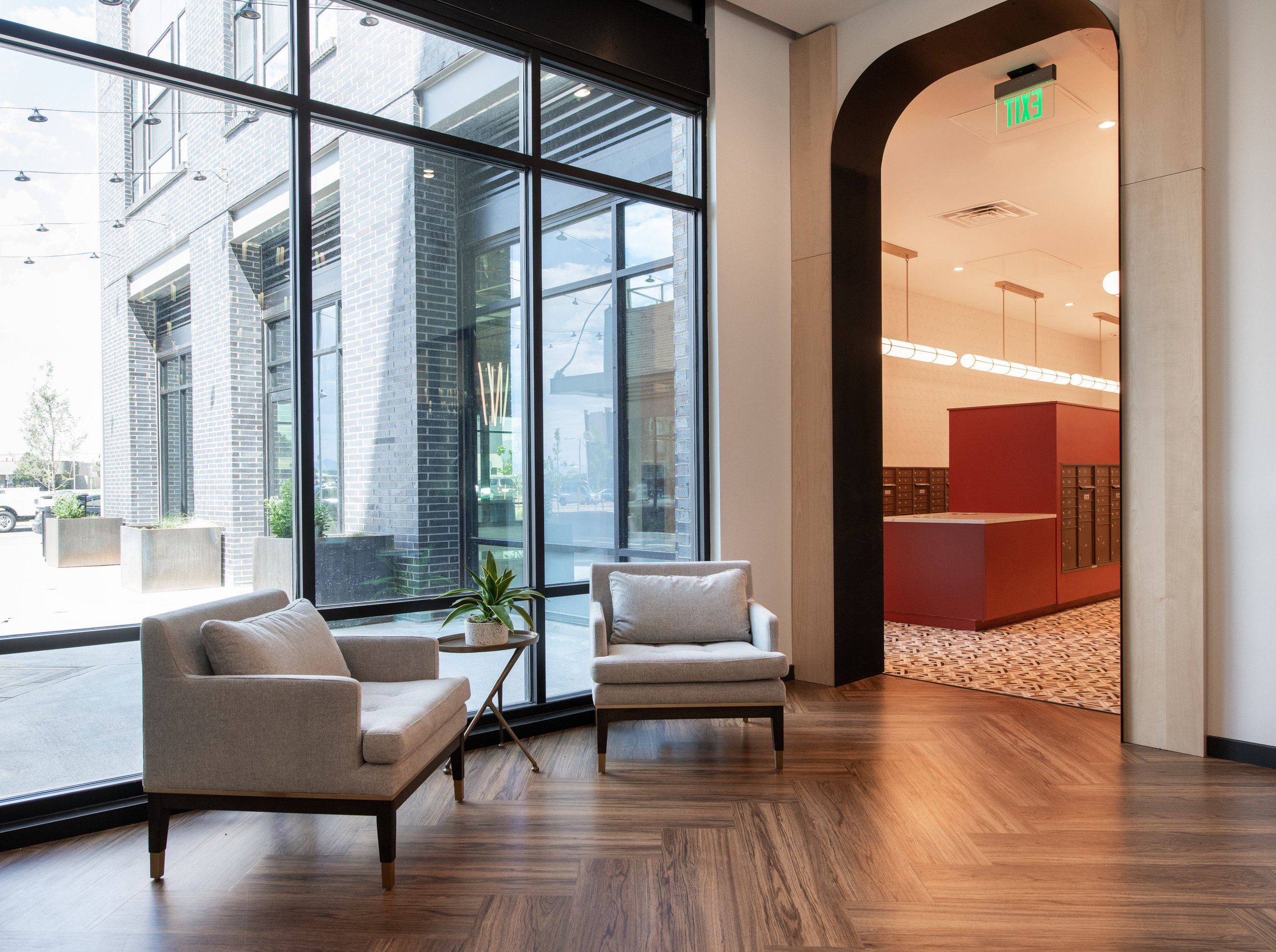 A modern lobby with large windows and two beige armchairs with a small coffee table and potted plant between them, adjacent to a hallway with wood flooring and mailboxes visible inside.