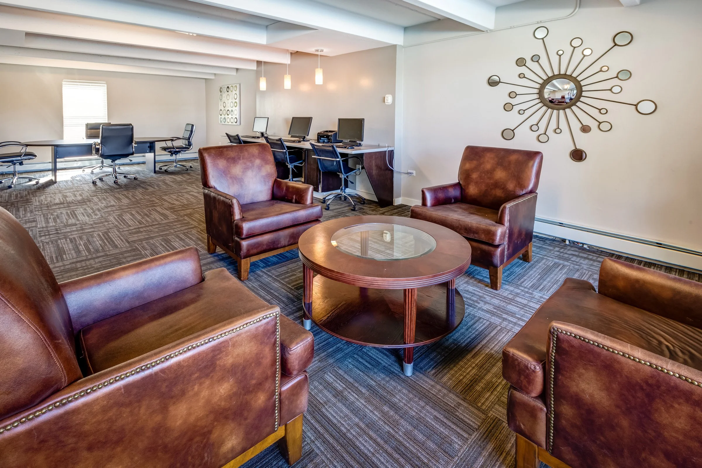 A lounge area with four brown leather armchairs around a round wooden coffee table with a glass top. In the background, there is a workspace with several desks, computers, and office chairs. A modern wall mirror decorates the light-colored wall, and 