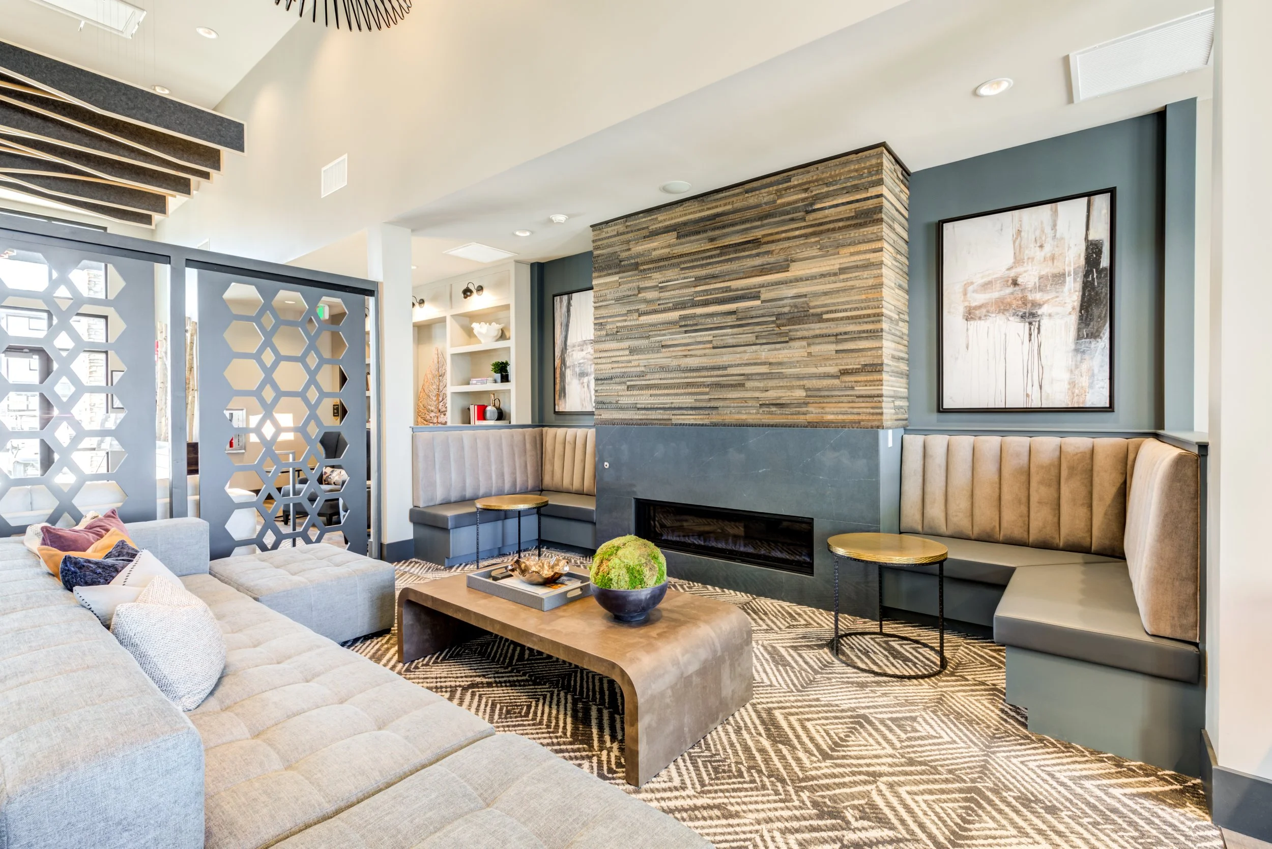 Modern living room with beige sectional sofa, patterned carpet, a rectangular wooden coffee table with decorative items including a plant, built-in beige and gray upholstered corner seating with small round tables, a fireplace with a wooden and stone