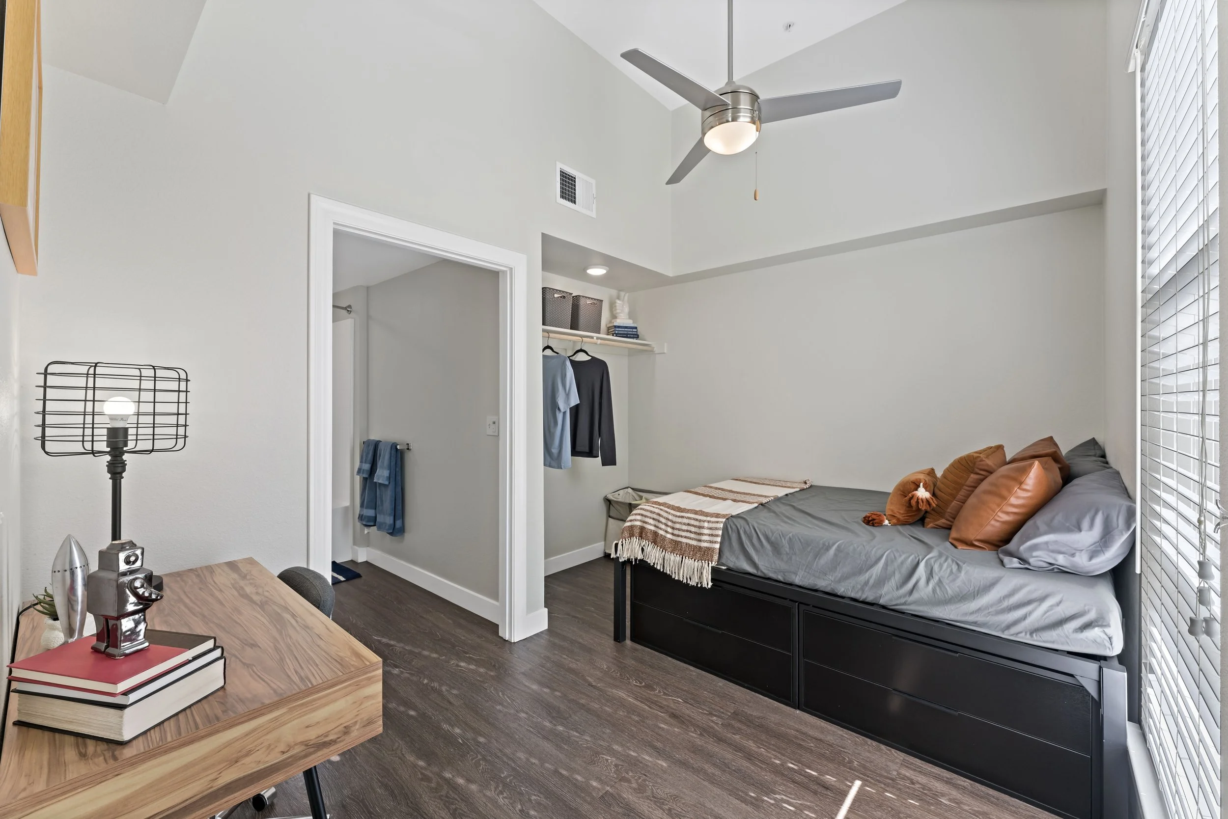 Small bedroom with a bed against the wall, decorative brown pillows, a blanket, a ceiling fan, a window with blinds, a wooden desk with books and a lamp, and an open closet with clothes and storage bins.