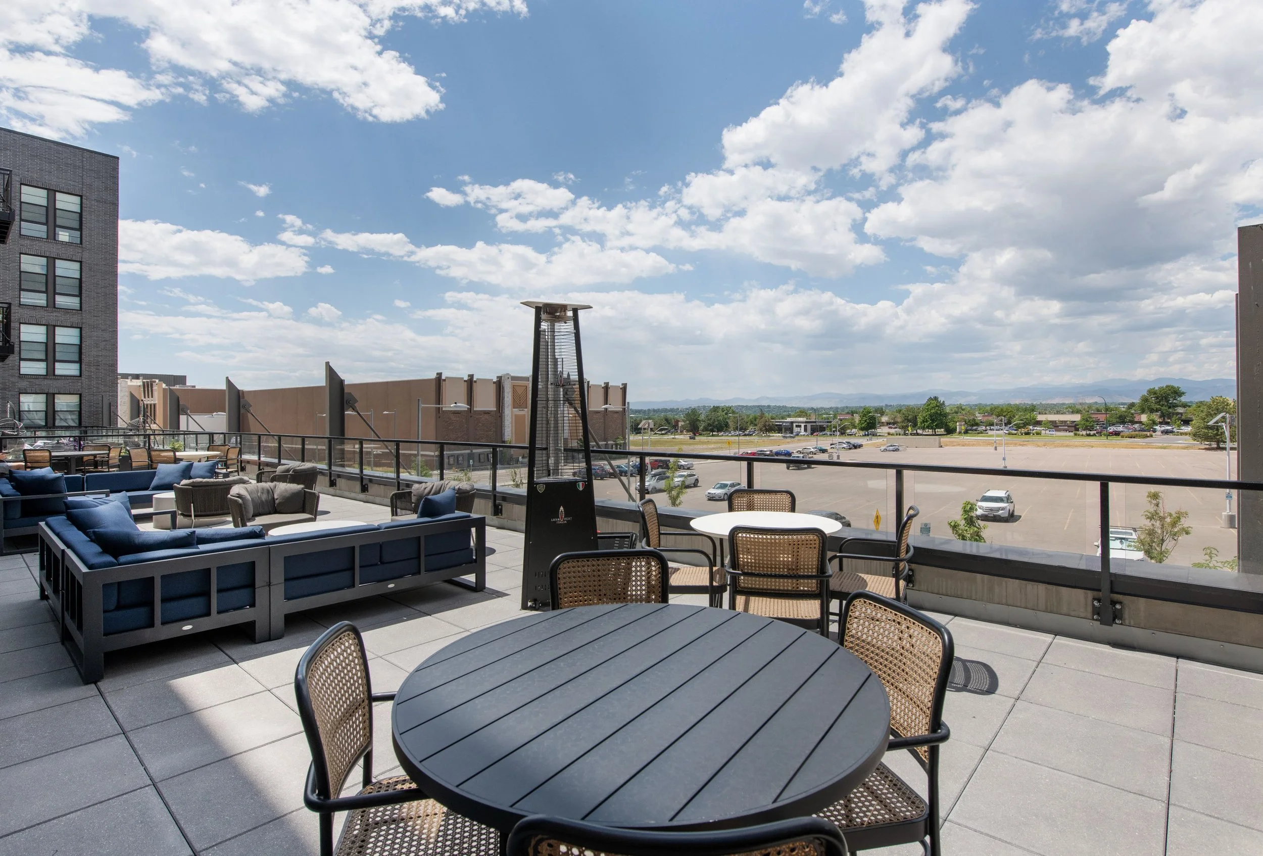 Outdoor rooftop patio with black round table and chairs, blue couches, and a tall patio heater, overlooking a parking lot and open landscape under partly cloudy sky.