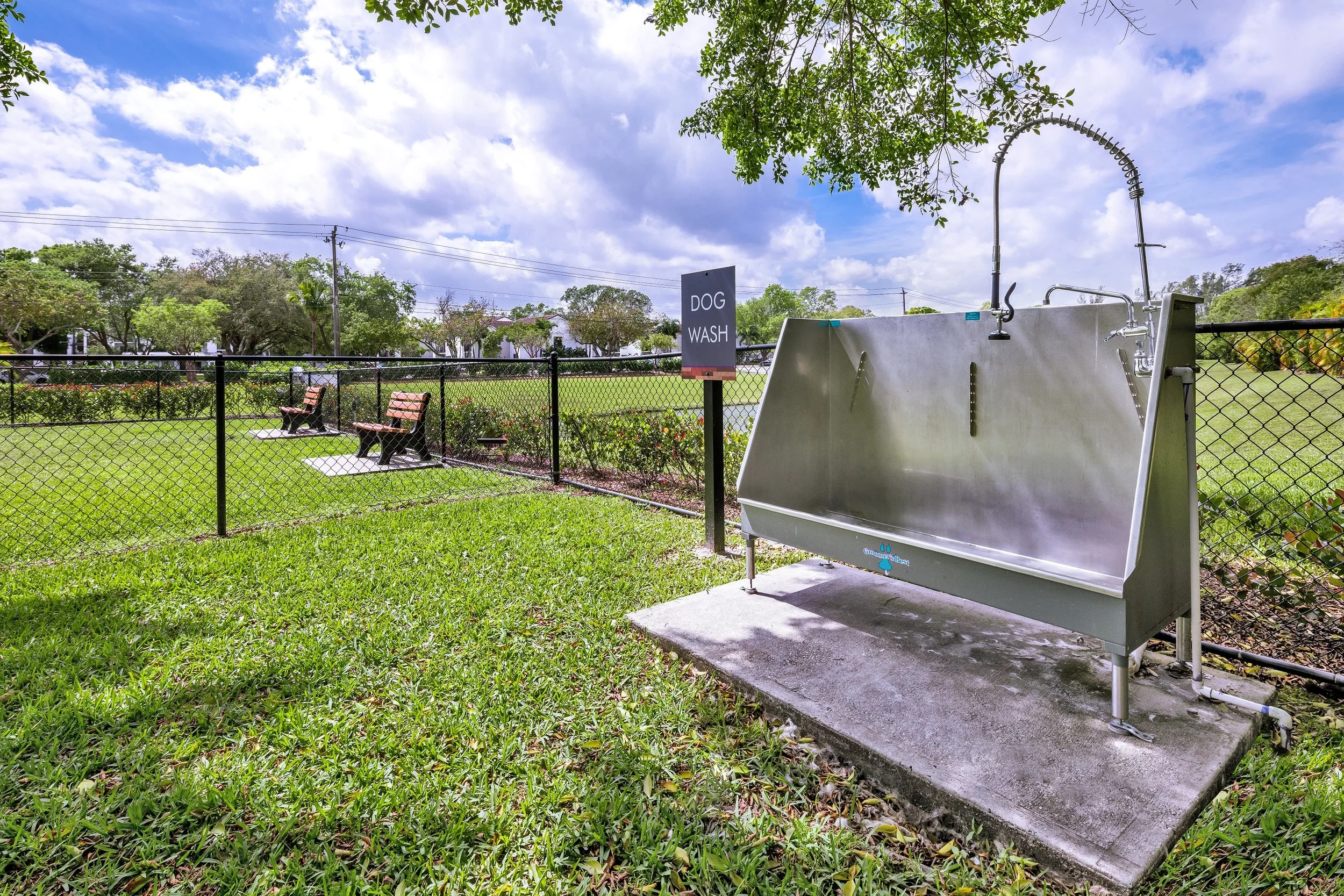 Outdoor dog wash station with a large metal tub and faucets, surrounded by a black fence and green grass. There are park benches in the background under a partly cloudy sky.