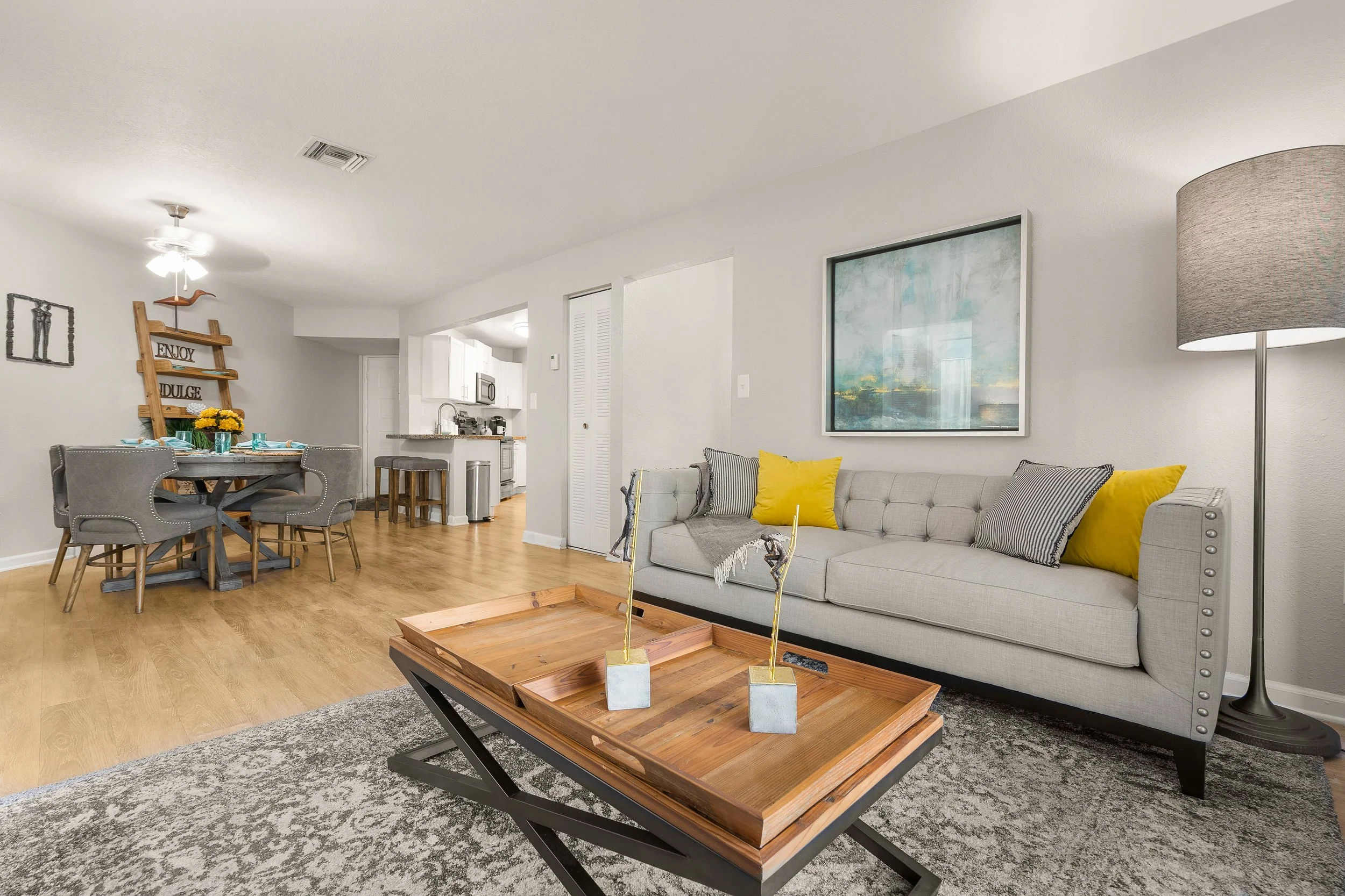Living room with beige sofa, yellow and striped cushions, wooden coffee table with decorative items, wall art, floor lamp, dining area with round table and six gray chairs, kitchen in background, hardwood floor, and neutral walls.