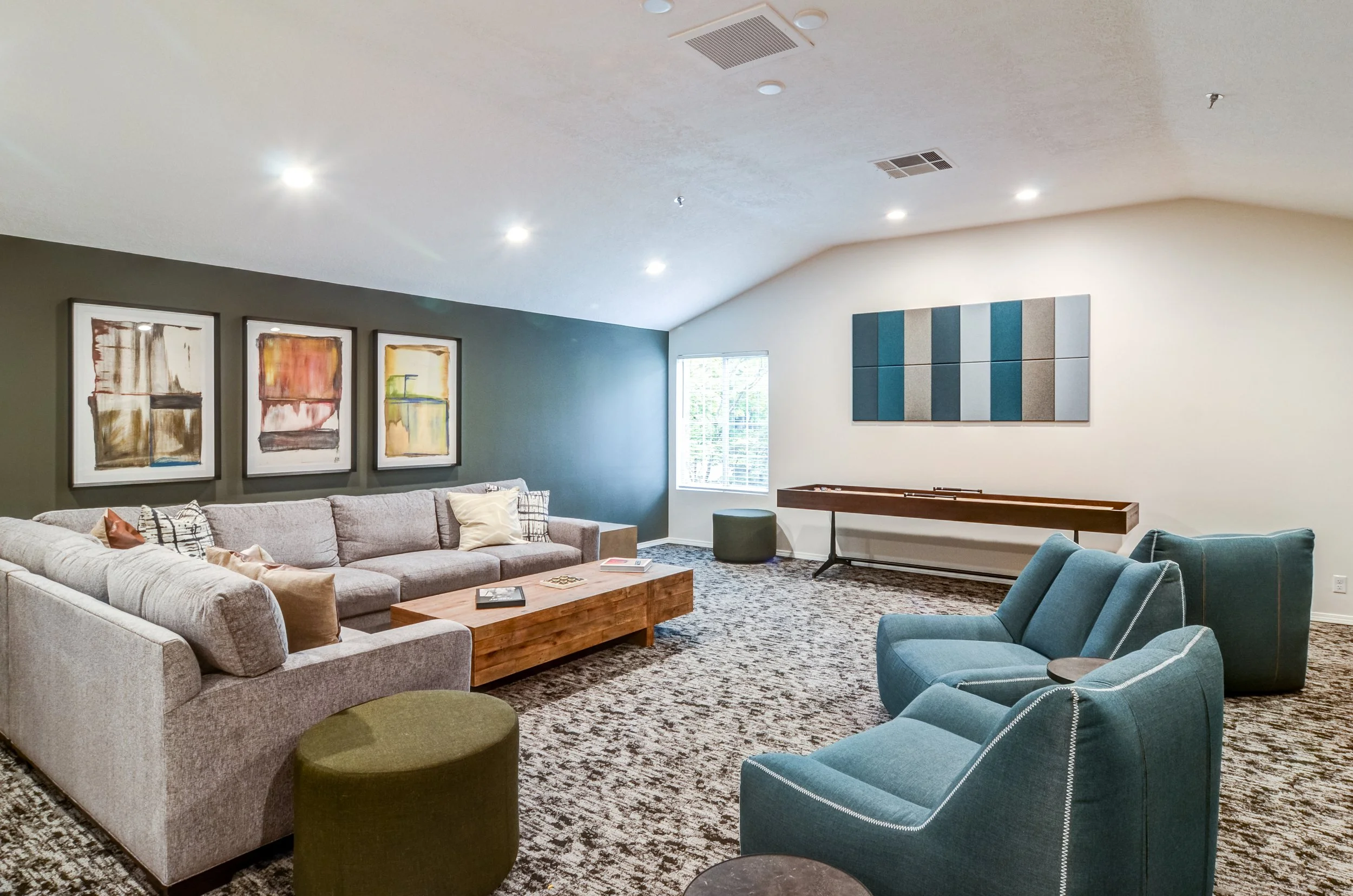 A living room with a large gray sectional sofa, two blue armchairs, abstract artwork on the walls, a wooden coffee table, and a window with white blinds.