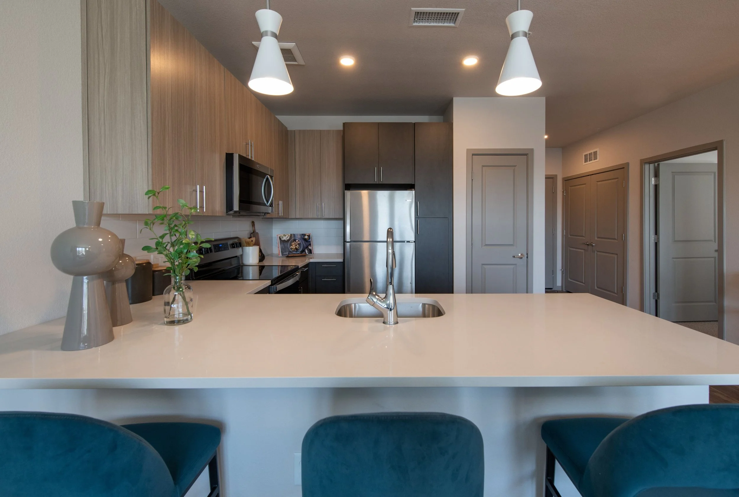 Modern kitchen with a white countertop island, a sink, a stainless steel fridge, microwave, oven, and cabinets in wood and dark gray finishes, with decorative vases and a glass of greenery on the counter.