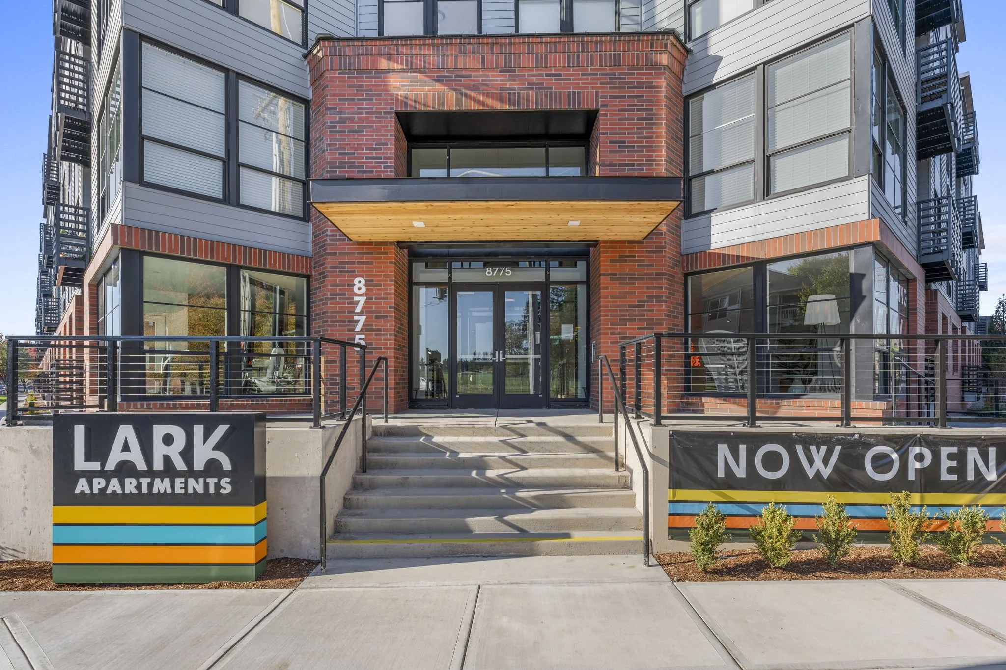 An apartment building with a modern brick and metal exterior. The entrance has glass doors, concrete steps, and a ramp. Signs indicate 'LARK APARTMENTS' and 'NOW OPEN'.