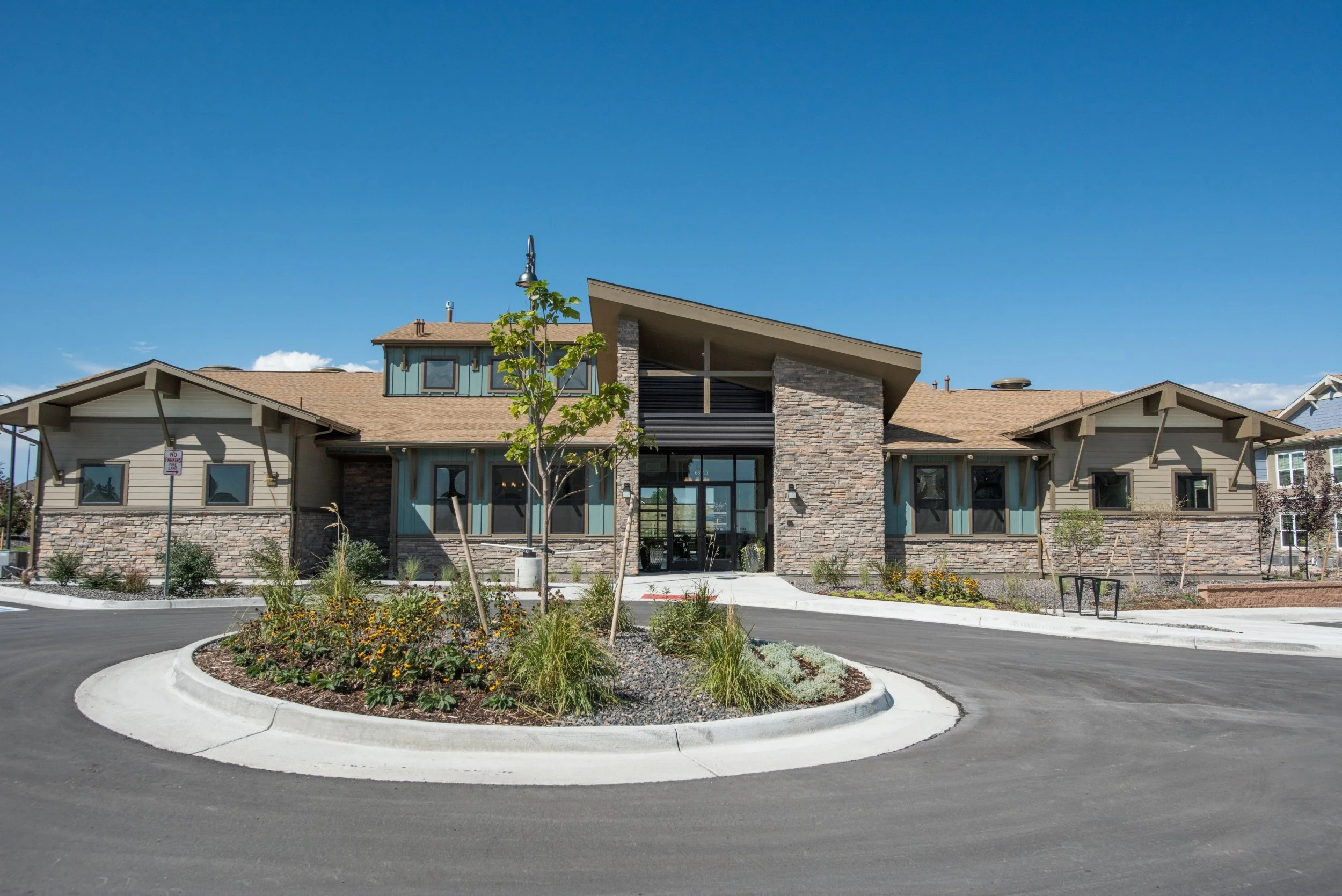 Modern residential building with stone and siding exterior, landscaped front yard, and a circular driveway under a clear blue sky.