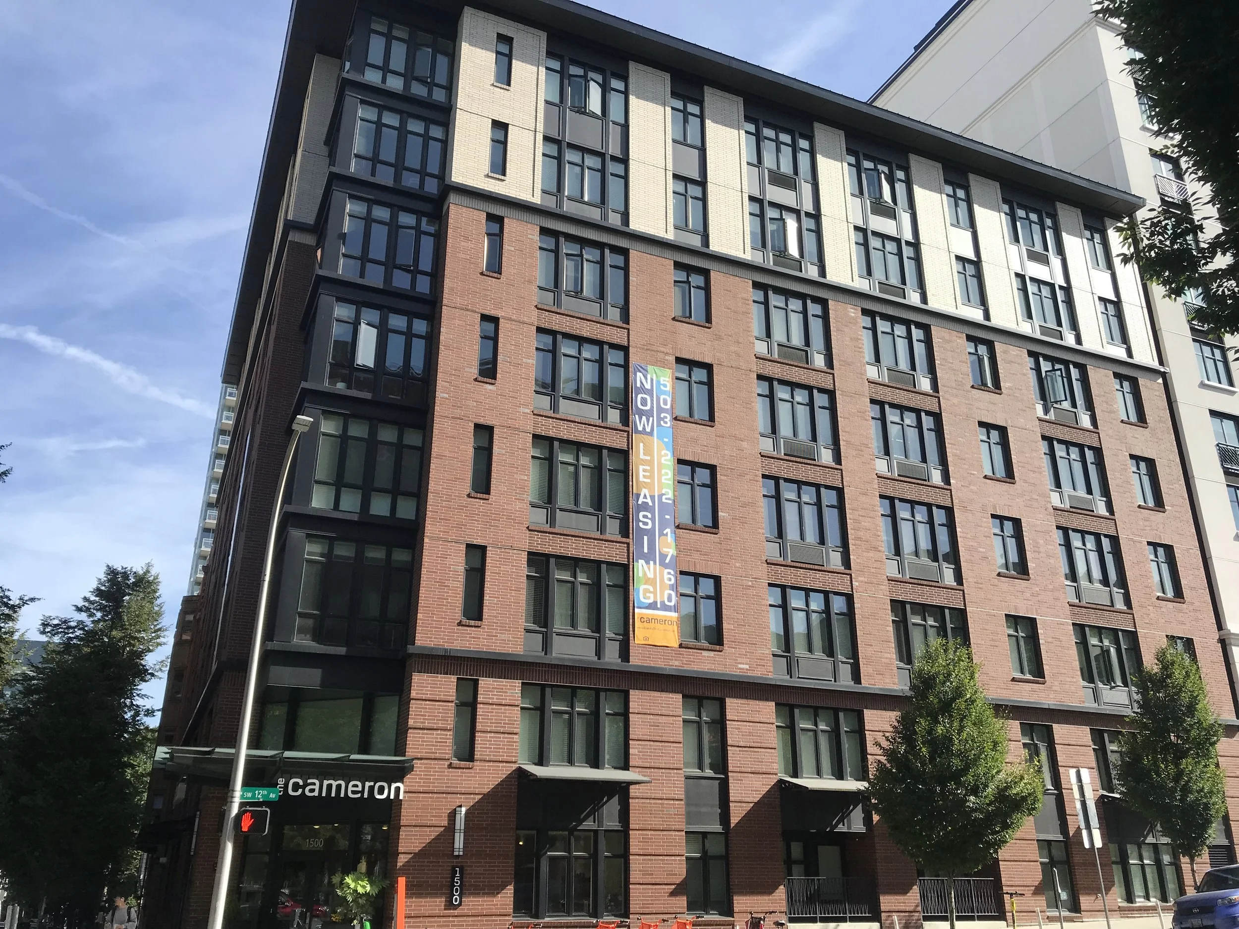 A multi-story modern apartment building with brick and dark metal siding, featuring numerous large windows. A tall banner on the building announces it as 'Now Leasing' with a contact number and address at the corner of the street. The street corner shows a traffic light, street sign, and some trees along the sidewalk.
