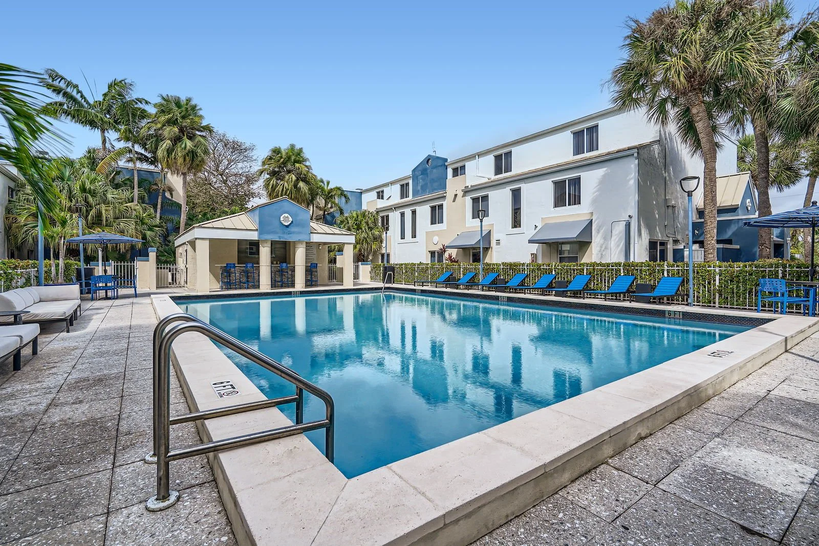 Empty outdoor swimming pool with blue lounge chairs, surrounded by palm trees and apartment buildings under clear sky.