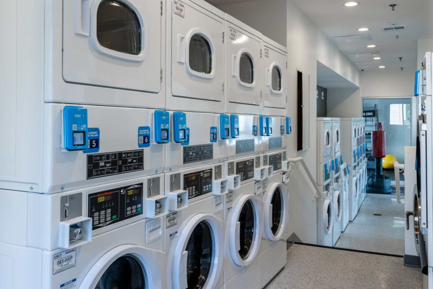 Laundry room with multiple front-loading washing machines and dryers, blue control panels, and a yellow exercise ball in the background.