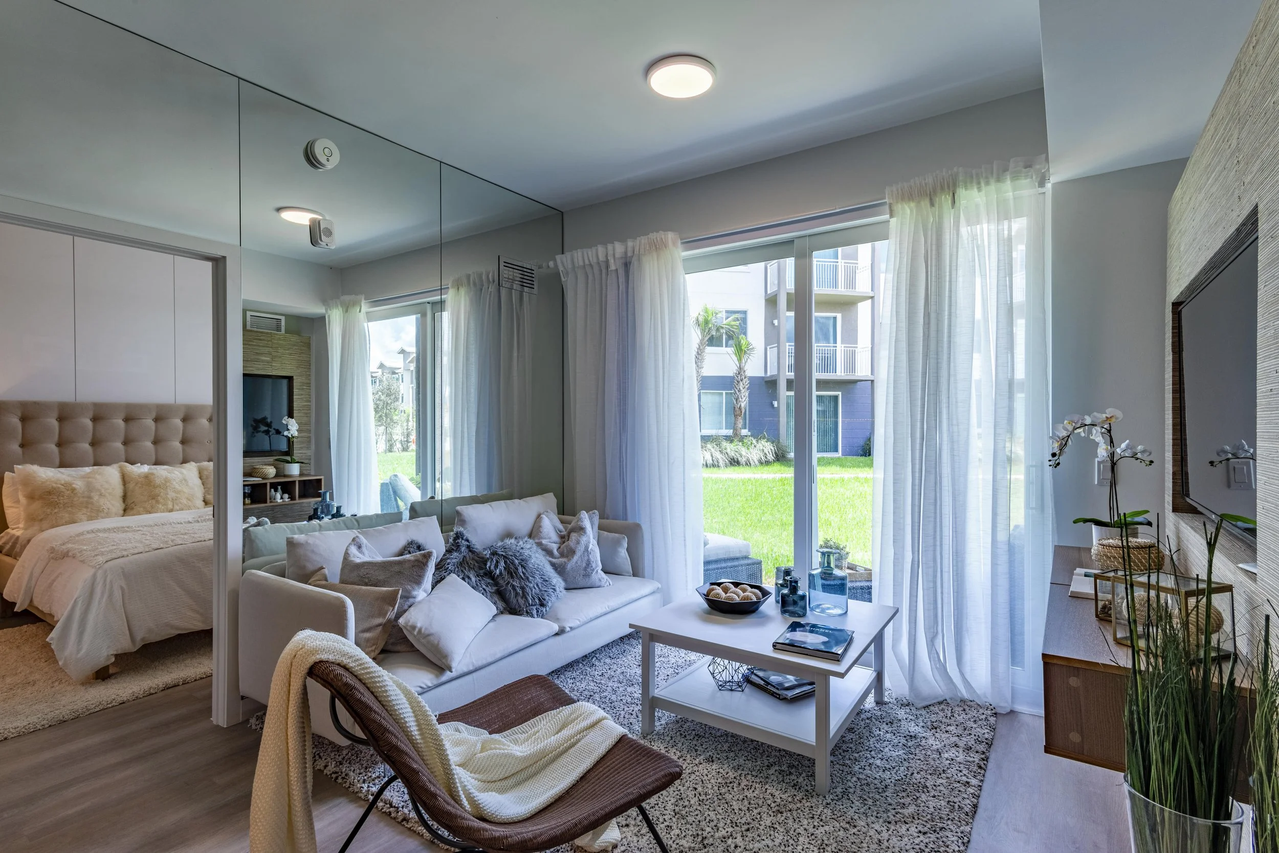 Living room with a white sofa, pillows, and a coffee table facing a large sliding glass door with sheer curtains, leading to a green outdoor space. Adjacent bedroom with a bed, headboard, and pillows visible through an open partition.