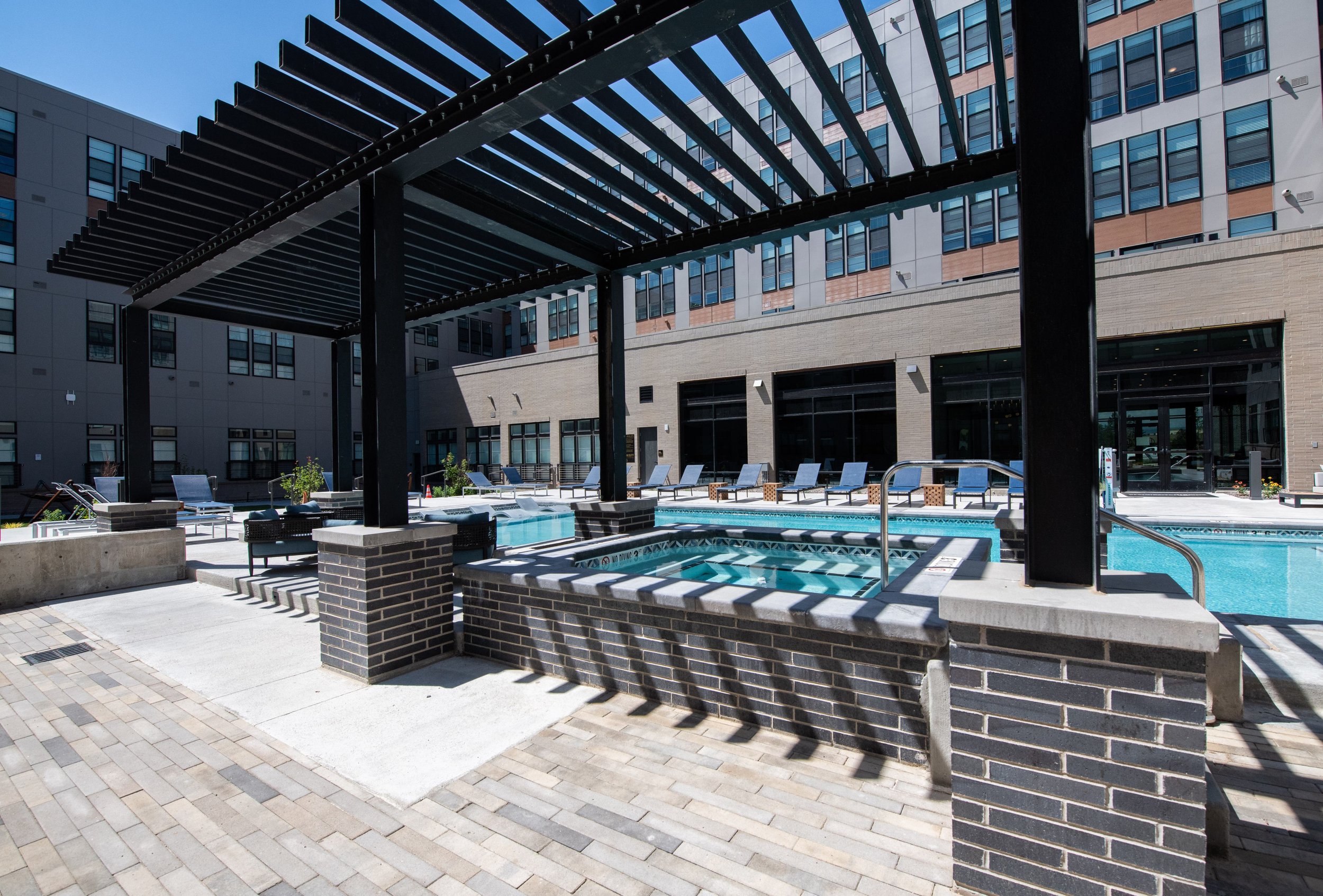 Outdoor swimming pool area with lounge chairs, a hot tub, and a pergola at a modern building complex.