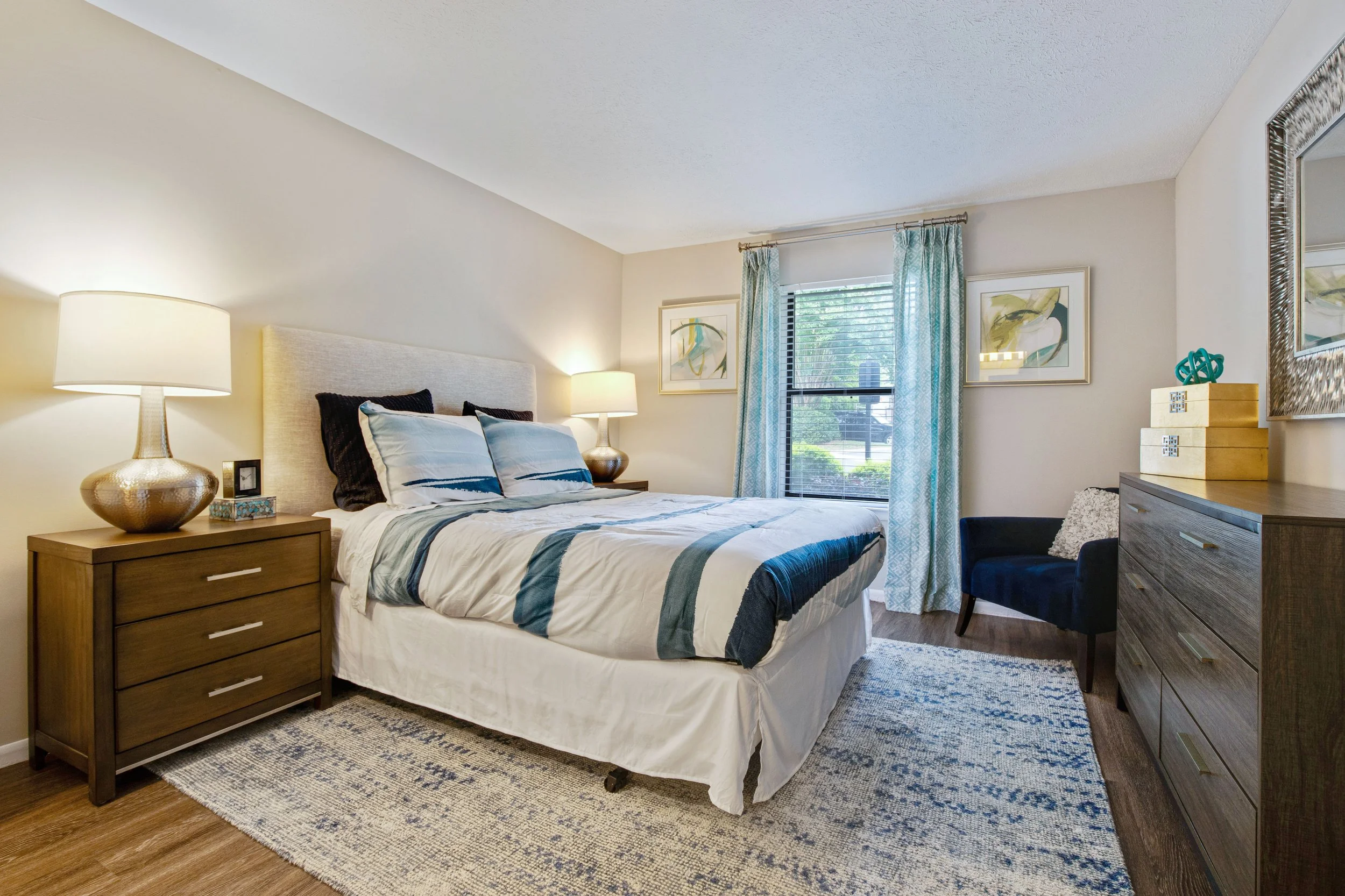 A cozy bedroom with a bed that has a upholstered headboard, striped bedding, and multiple pillows. On the left, a wooden nightstand with a large table lamp and small decorative item. To the right, a dresser with decor and a mirror above it. Light fil