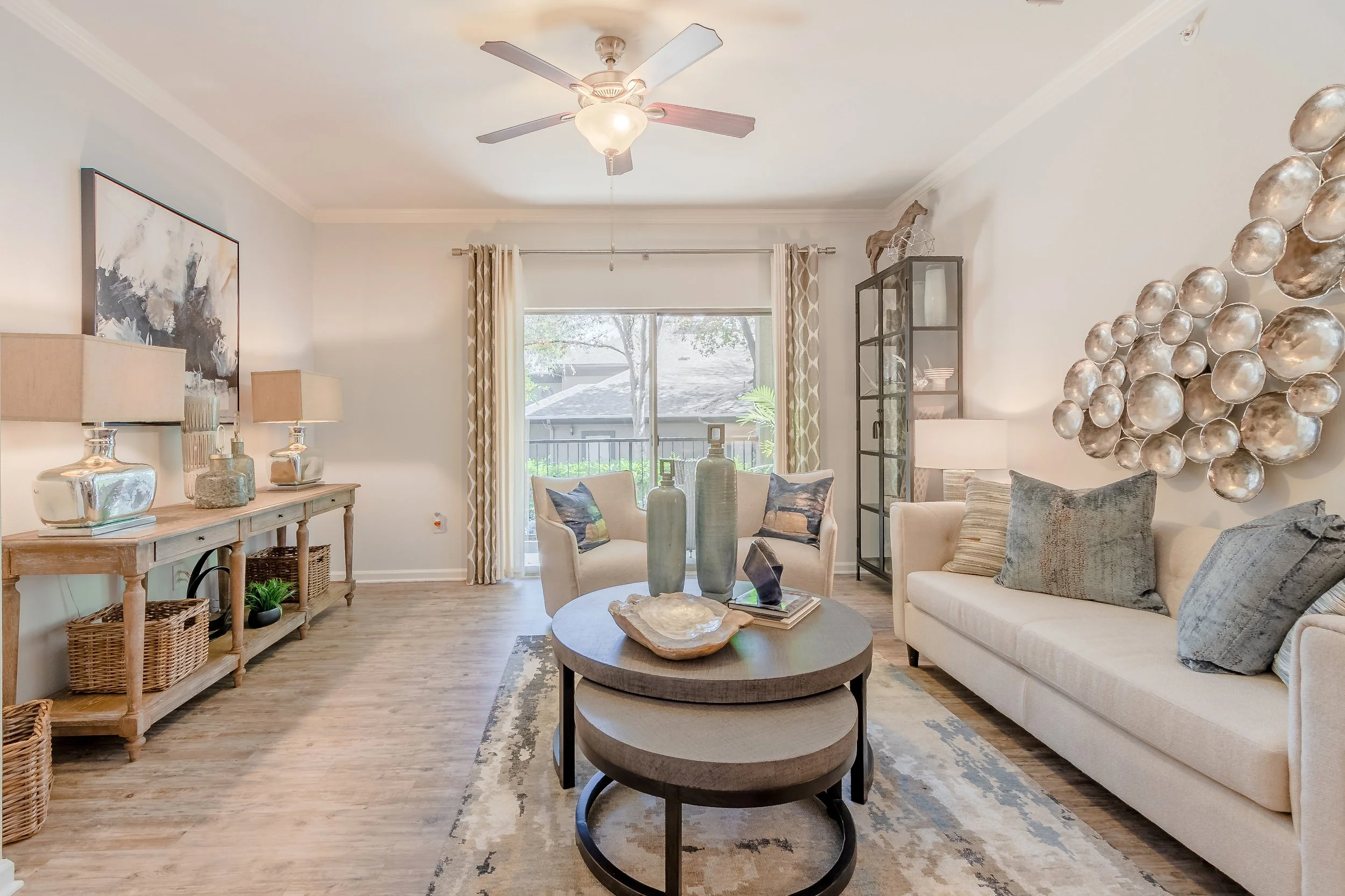 Living room with a beige sofa and two armchairs, a round coffee table with decorative vases and books, a wooden sideboard with lamps and decorative objects, a tall glass cabinet, a large metal wall art piece, hardwood floors, and curtains on a slidin