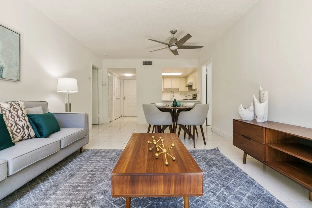 Living room with gray sofa, green and patterned throw pillows, wooden coffee table with decorative gold sculpture, gray rug, white walls, white ceiling fan, and a wooden TV stand with white vases, with a dining area and kitchen in the background.