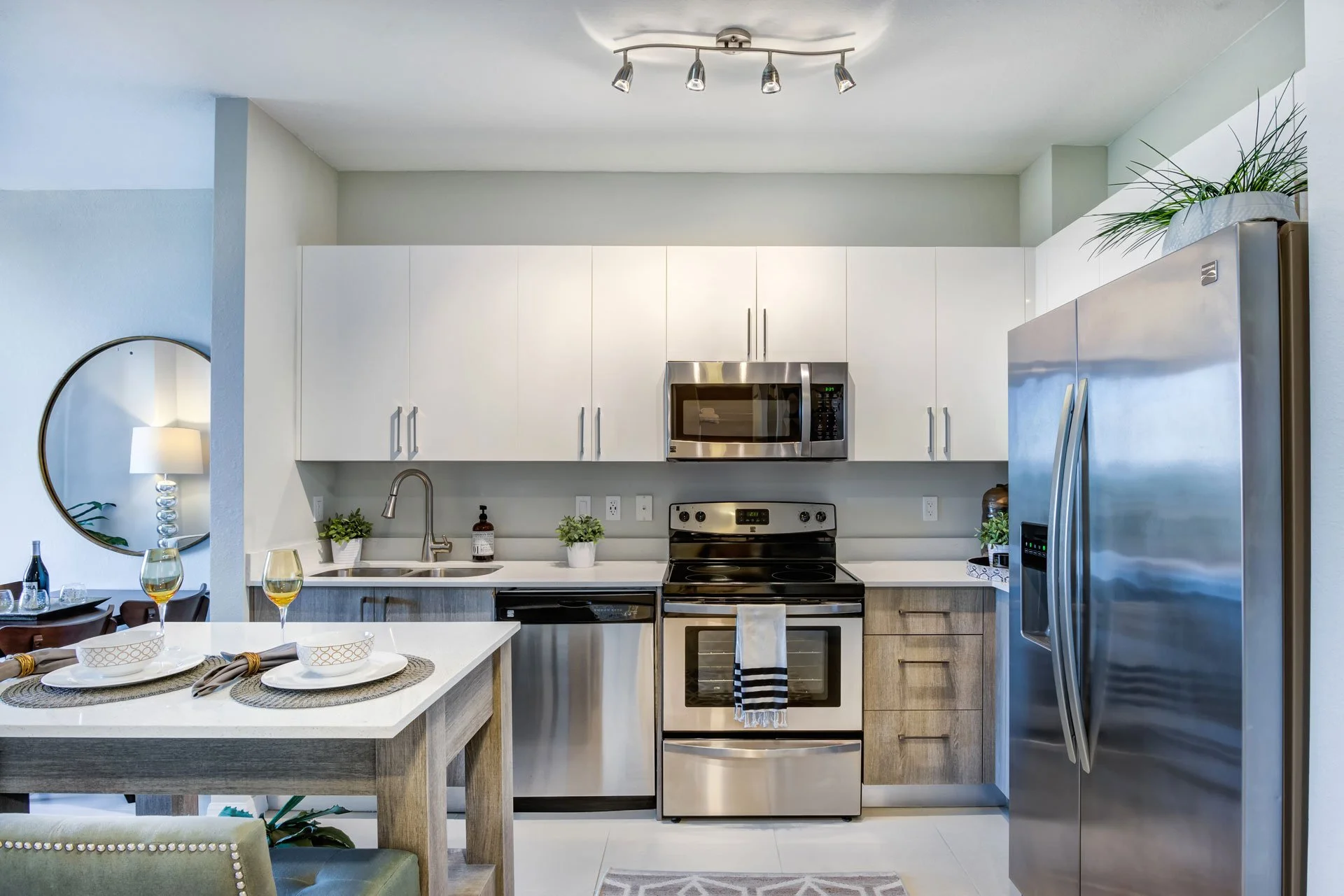 Modern kitchen with white cabinets, stainless steel appliances, a small dining table with place settings, and potted plants, illuminated by track lighting and natural light.