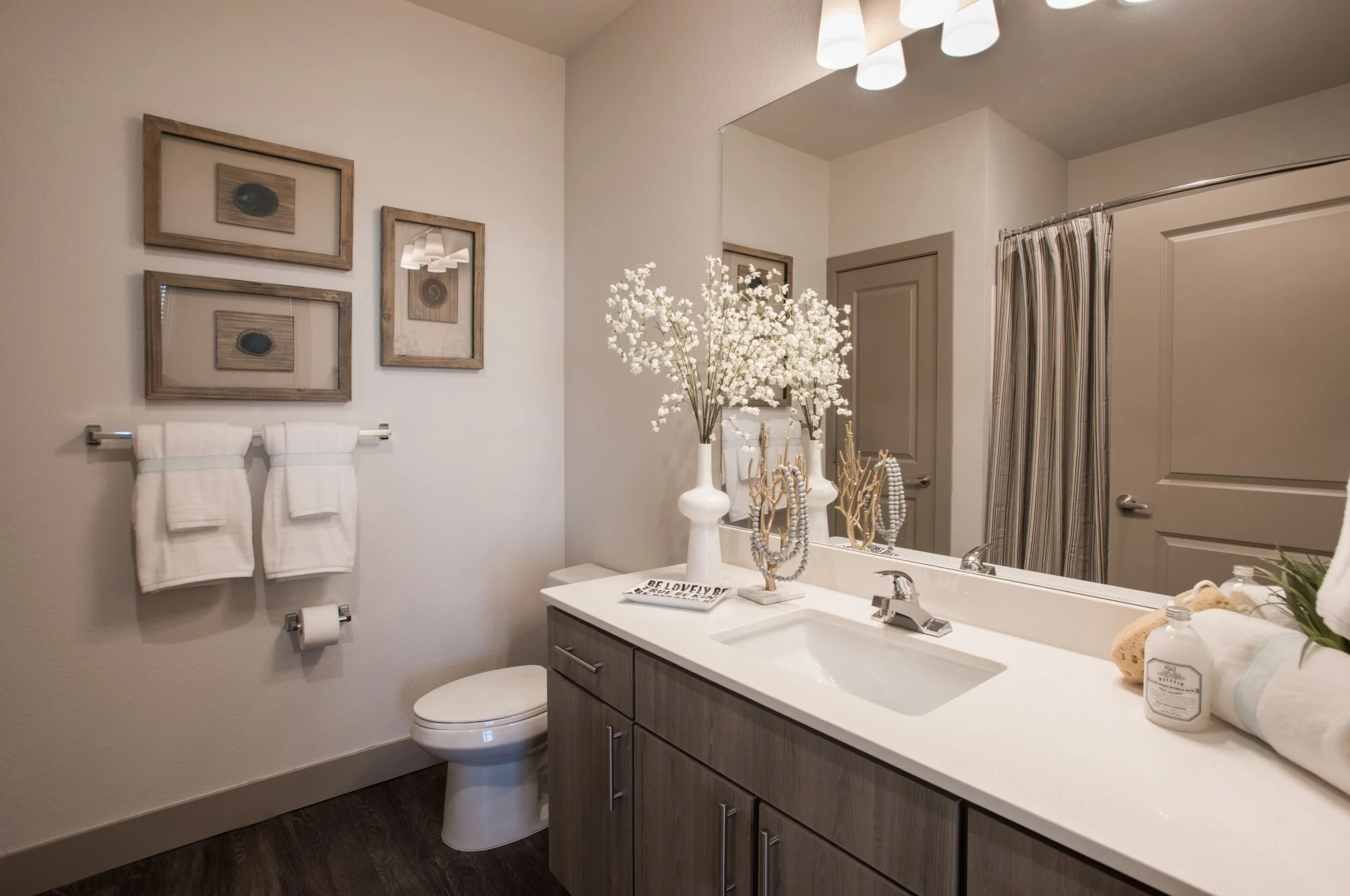 A bathroom with a vanity, large mirror, and decorative items including a large white vase with white flowers, framed artwork on the wall, and towels.