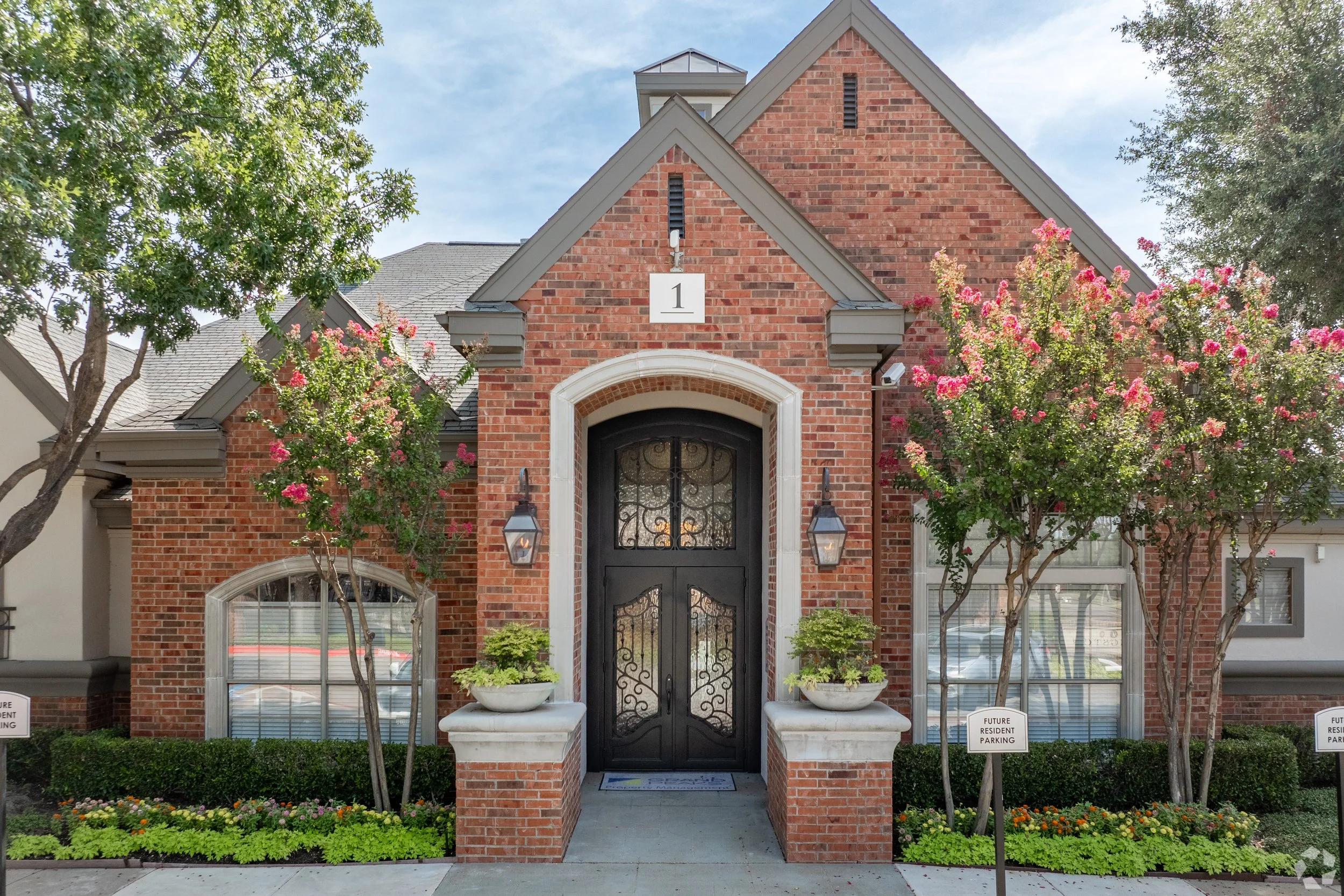 Front entrance of a brick residential building with a black wrought iron door, flanked by two potted plants, and pink flowering trees on both sides.