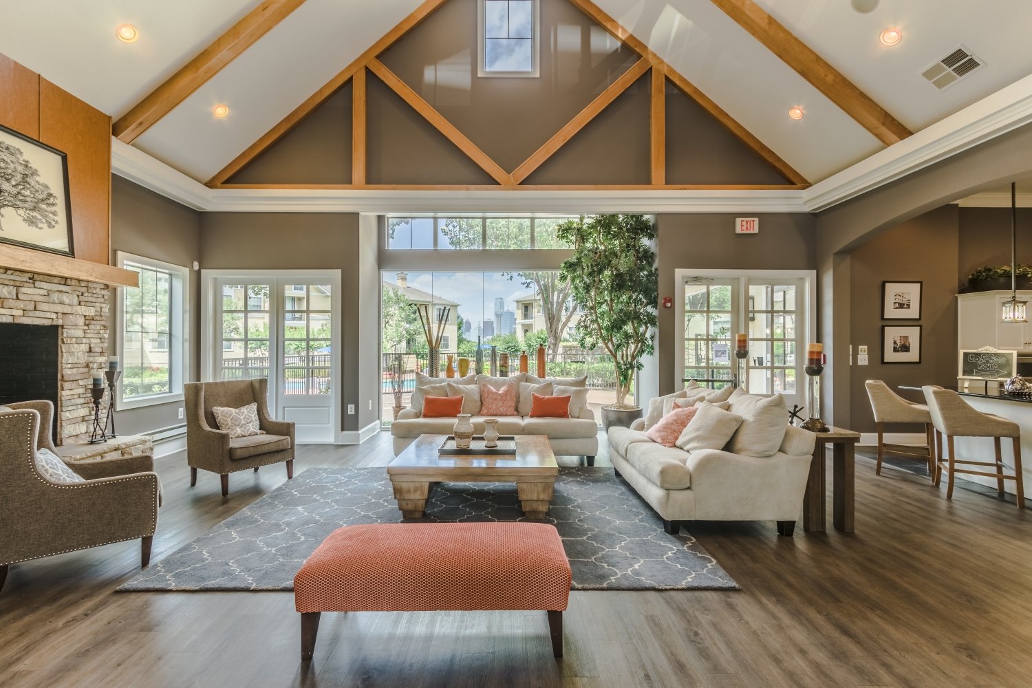 Spacious living room with high vaulted ceiling and wooden beams, large windows and glass doors providing natural light, comfortable sofas and armchairs, a stone fireplace, a coffee table, potted plants, and outdoor views of trees and city skyline