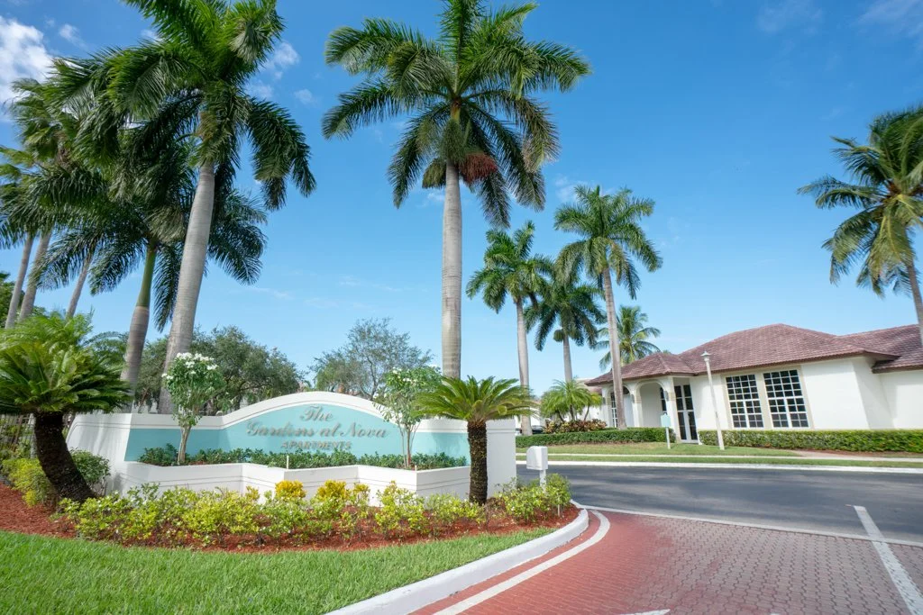 Entrance sign for The Cascades at Nona, a residential community with palm trees, a single-story house with a red-tiled roof, and a paved road and sidewalk.